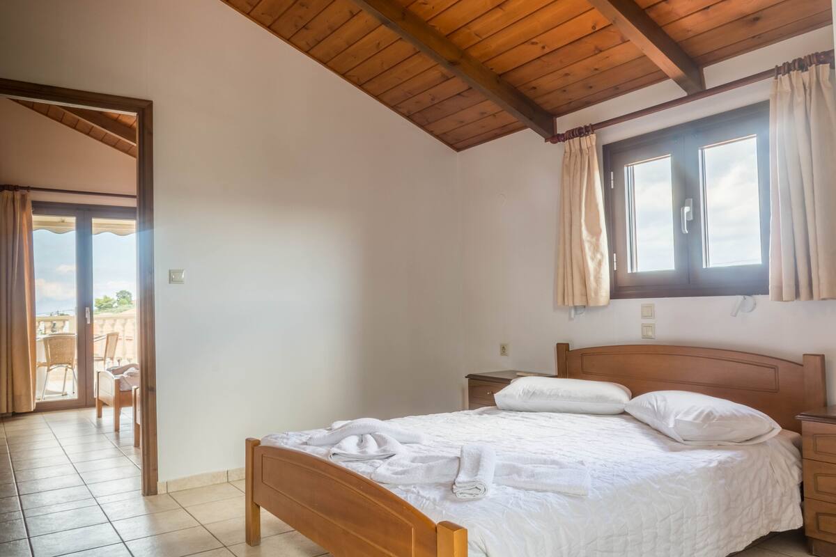 A bedroom with a wooden bed, a nightstand, a window with curtains, and a door leading to an outdoor area.