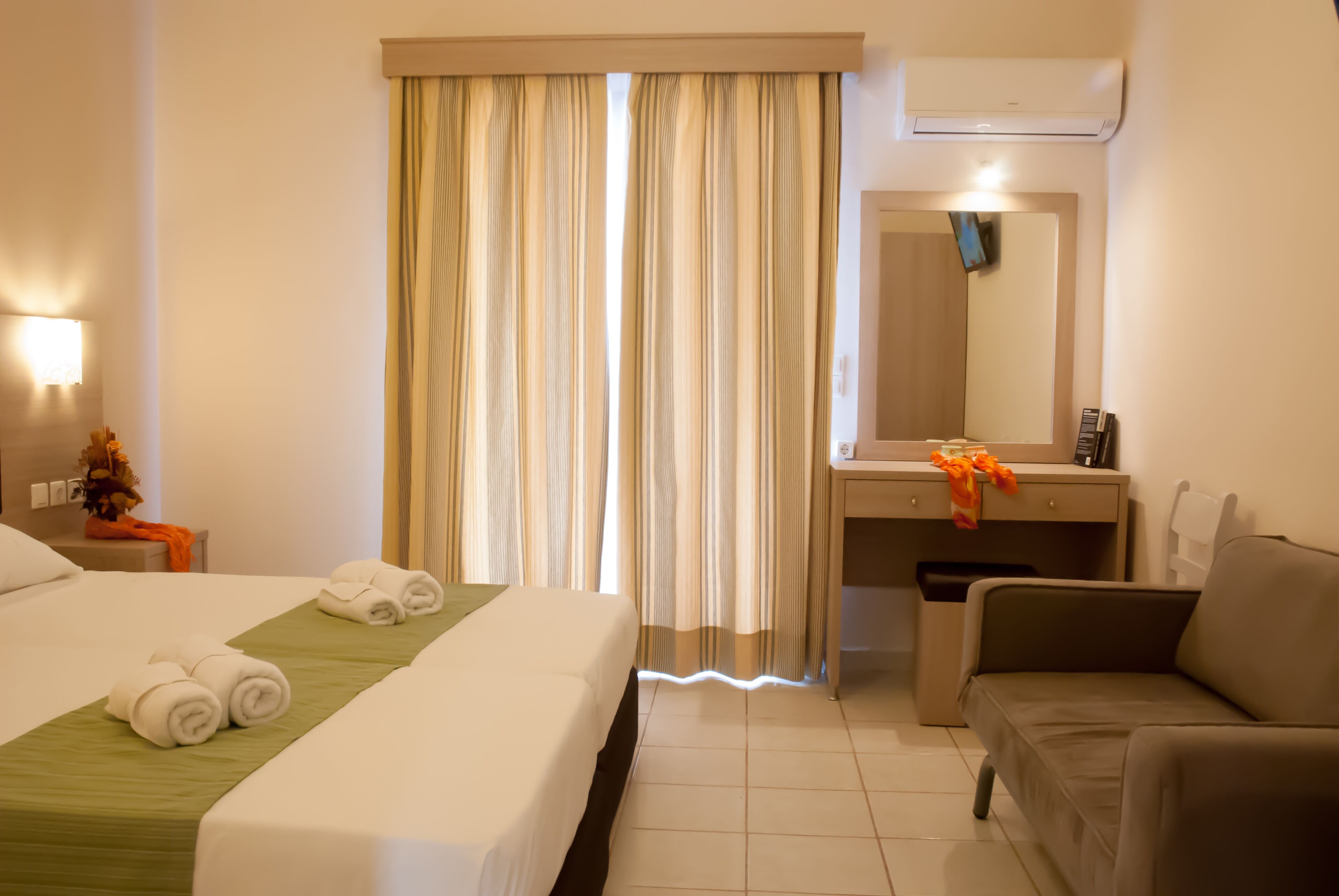 Quarto de hotel com cama, escrivaninha, cadeira e uma janela grande com cortinas.