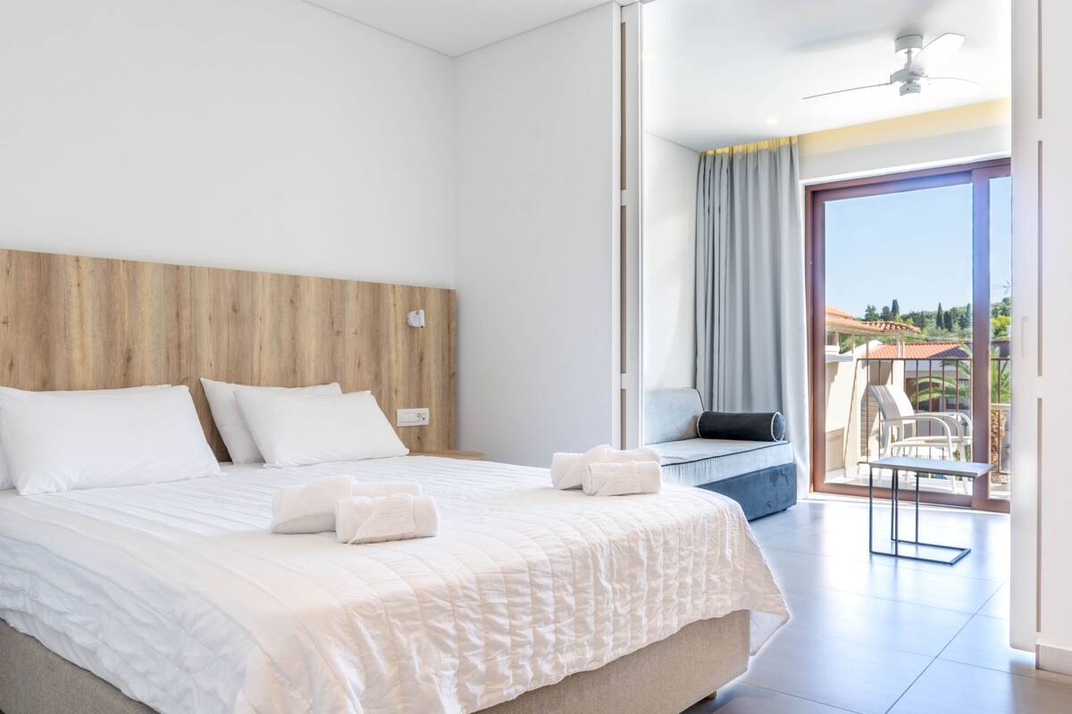 A modern hotel room with a large bed, a wooden headboard, a balcony with a view, and a ceiling fan.
