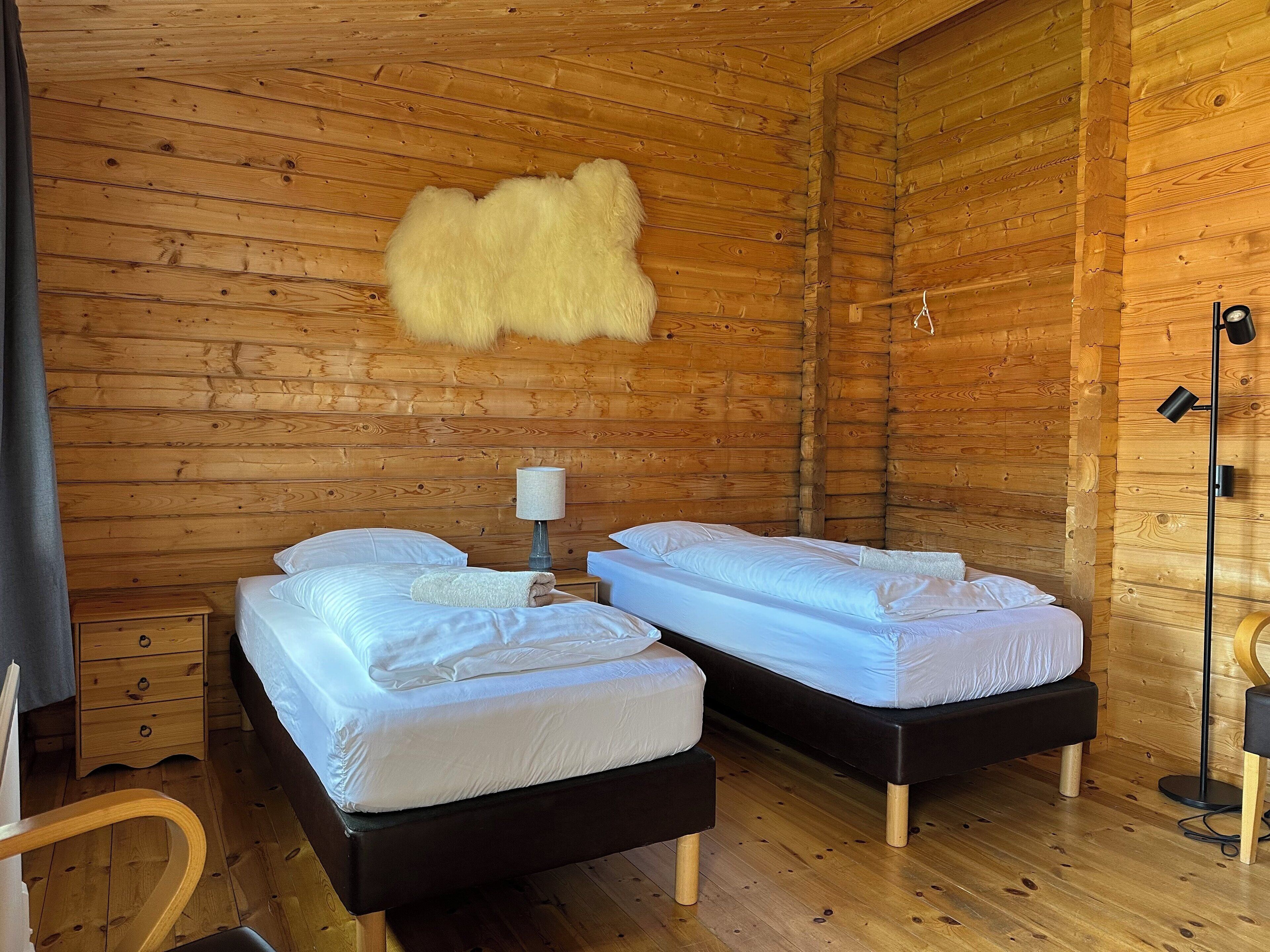 Una cabaña con dos camas, piso de madera, alfombra de piel y una lámpara.