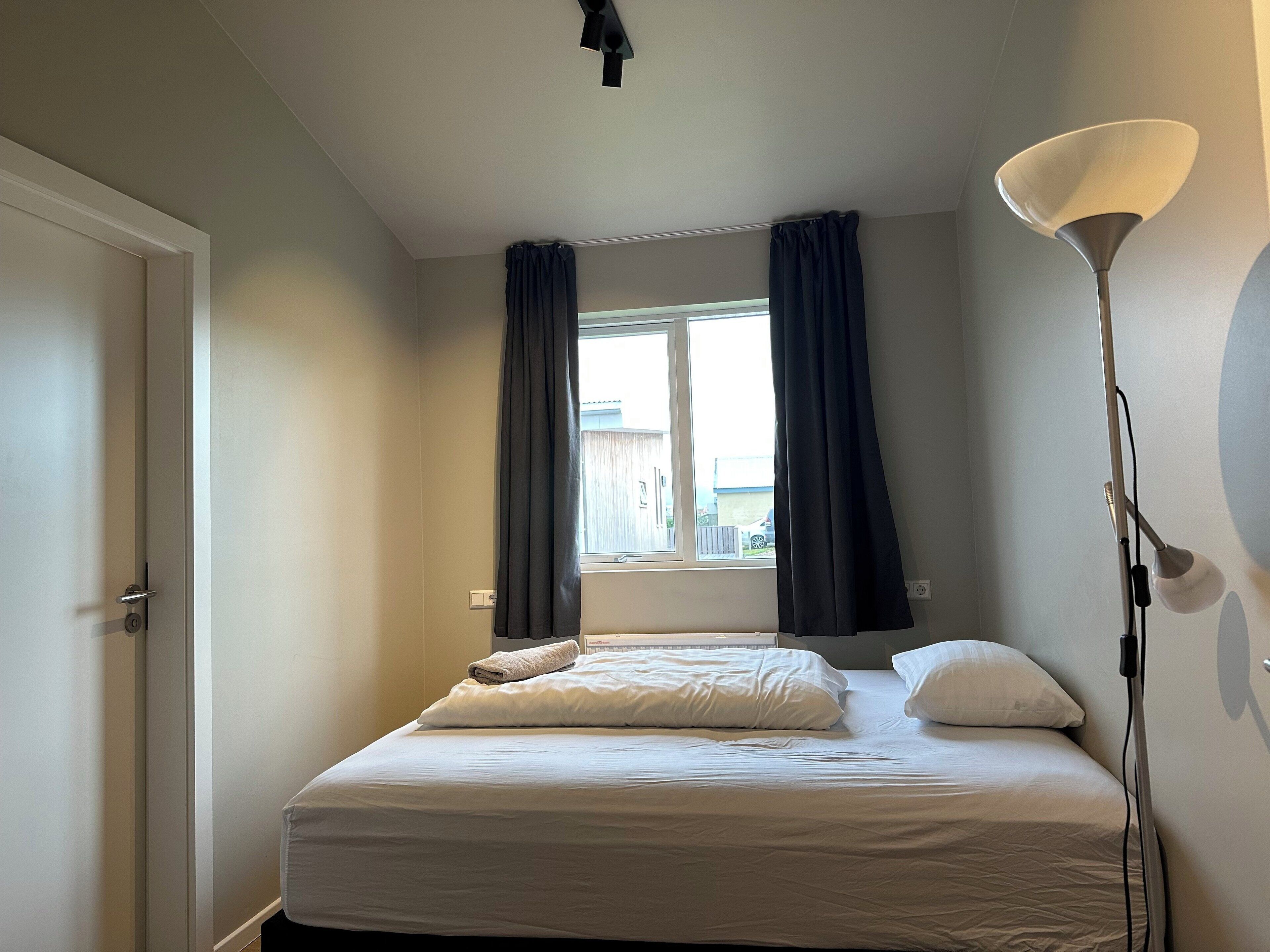 Un dormitorio con una cama, una ventana con cortinas, una lámpara de pie y una puerta.