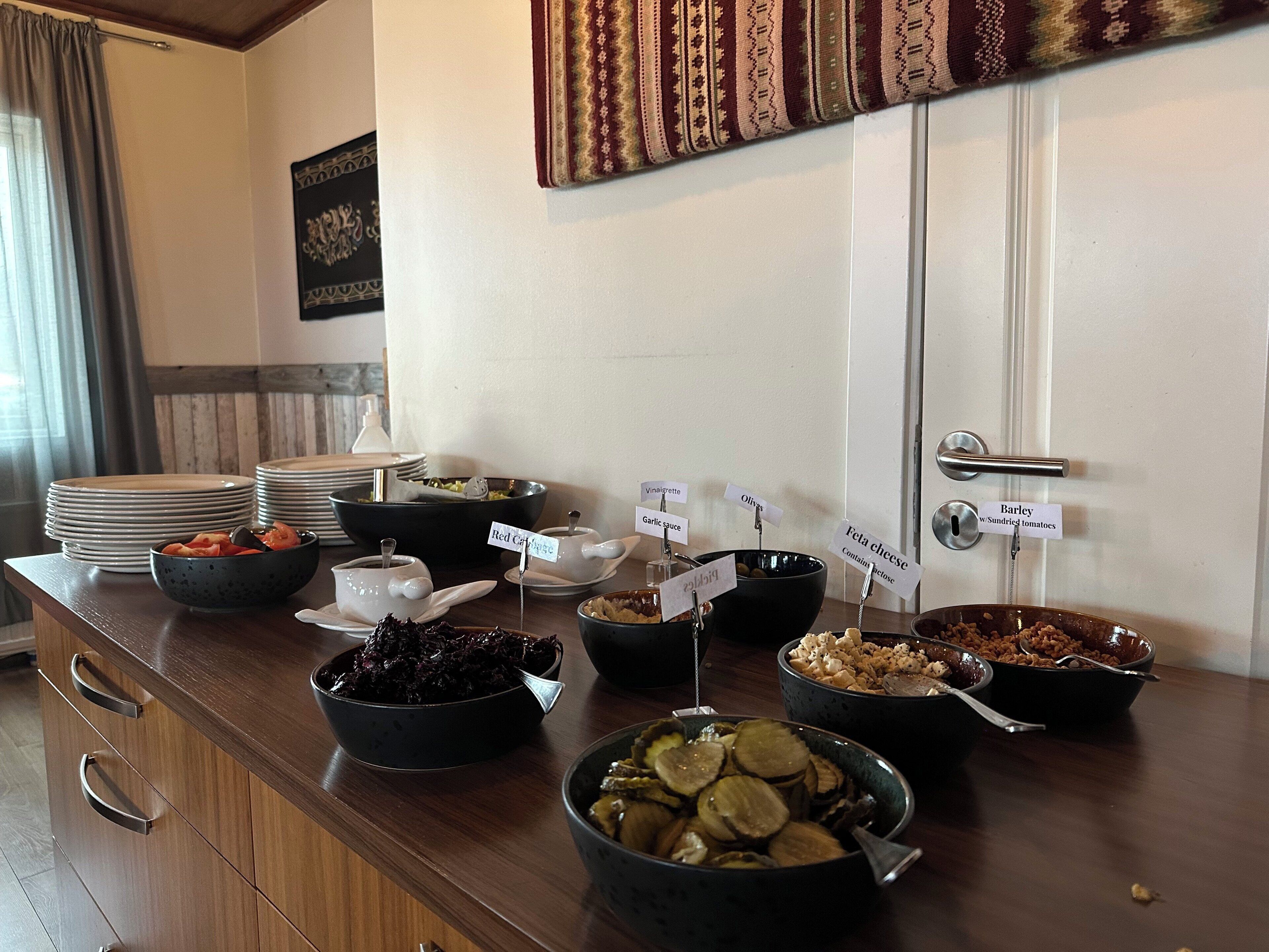 Una mesa de buffet con diversos alimentos y condimentos, incluyendo ensaladas, pepinillos y un recipiente con col roja.