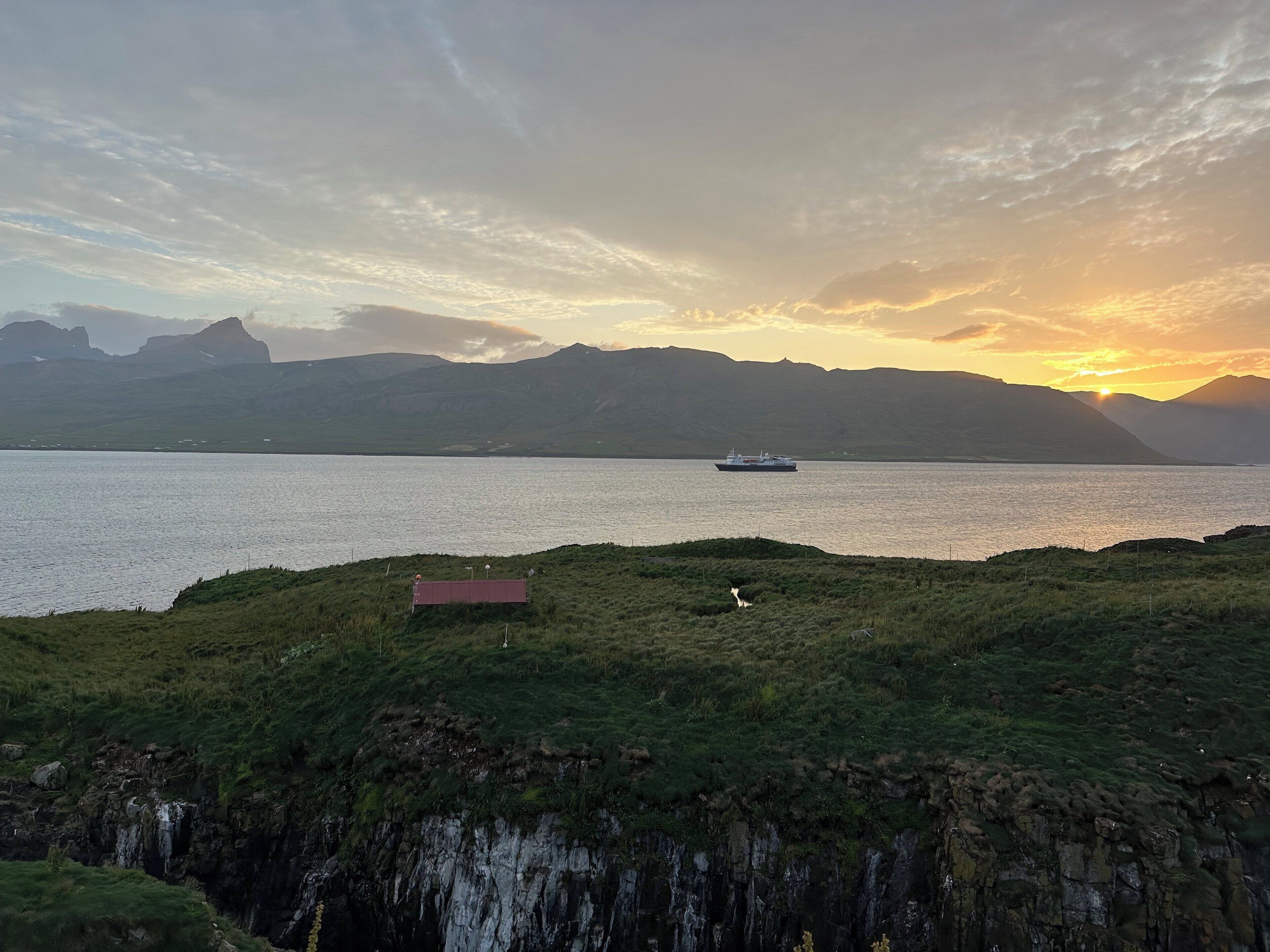 A coastal landscape with a ship on the water, a small building, and mountains in the background.