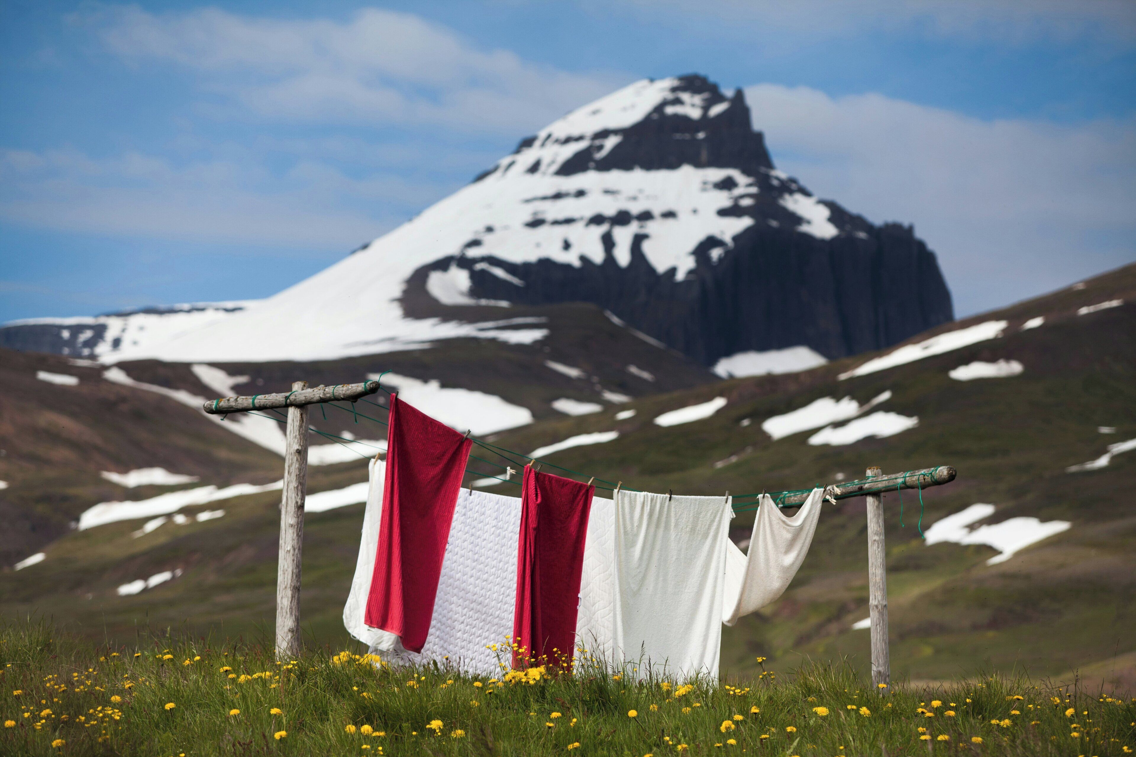 Una montaña nevada al fondo, un tendedero de madera con telas rojas y blancas, y un campo de flores amarillas en primer plano.
