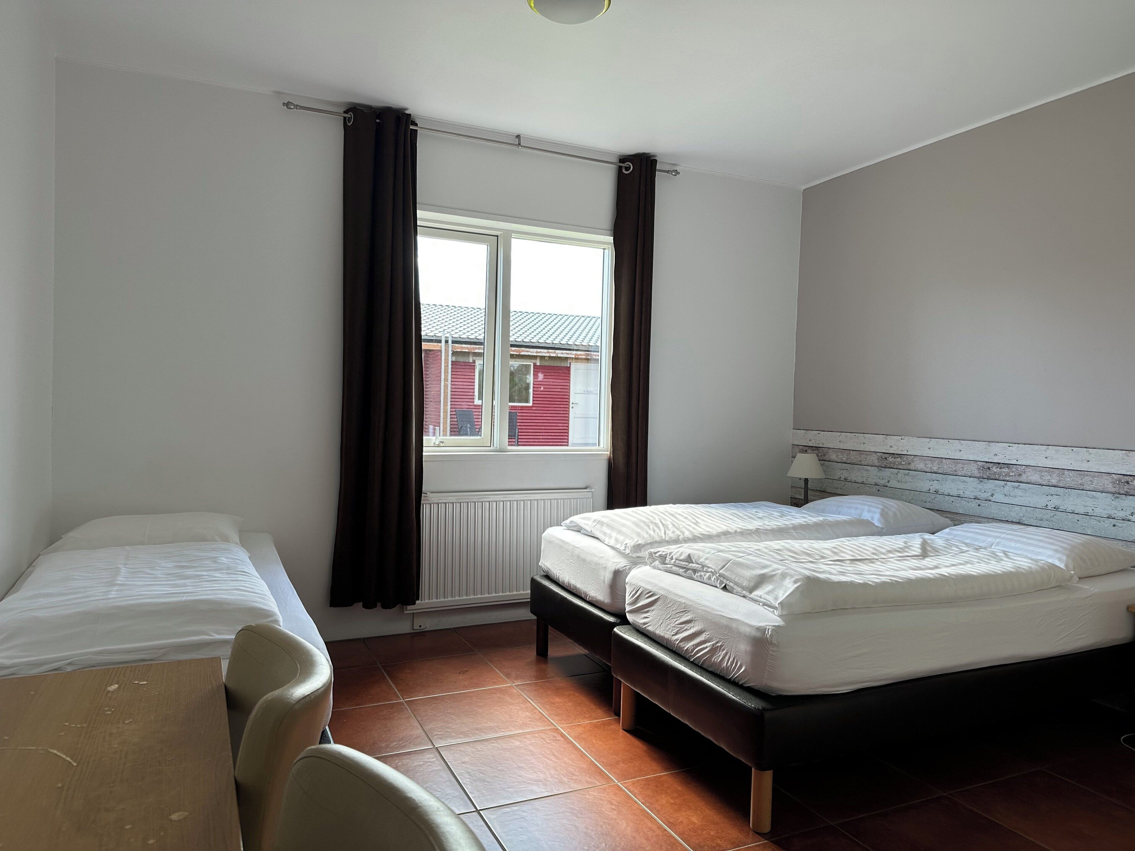 A bedroom with two beds, a window with curtains, a radiator, and a wooden headboard.