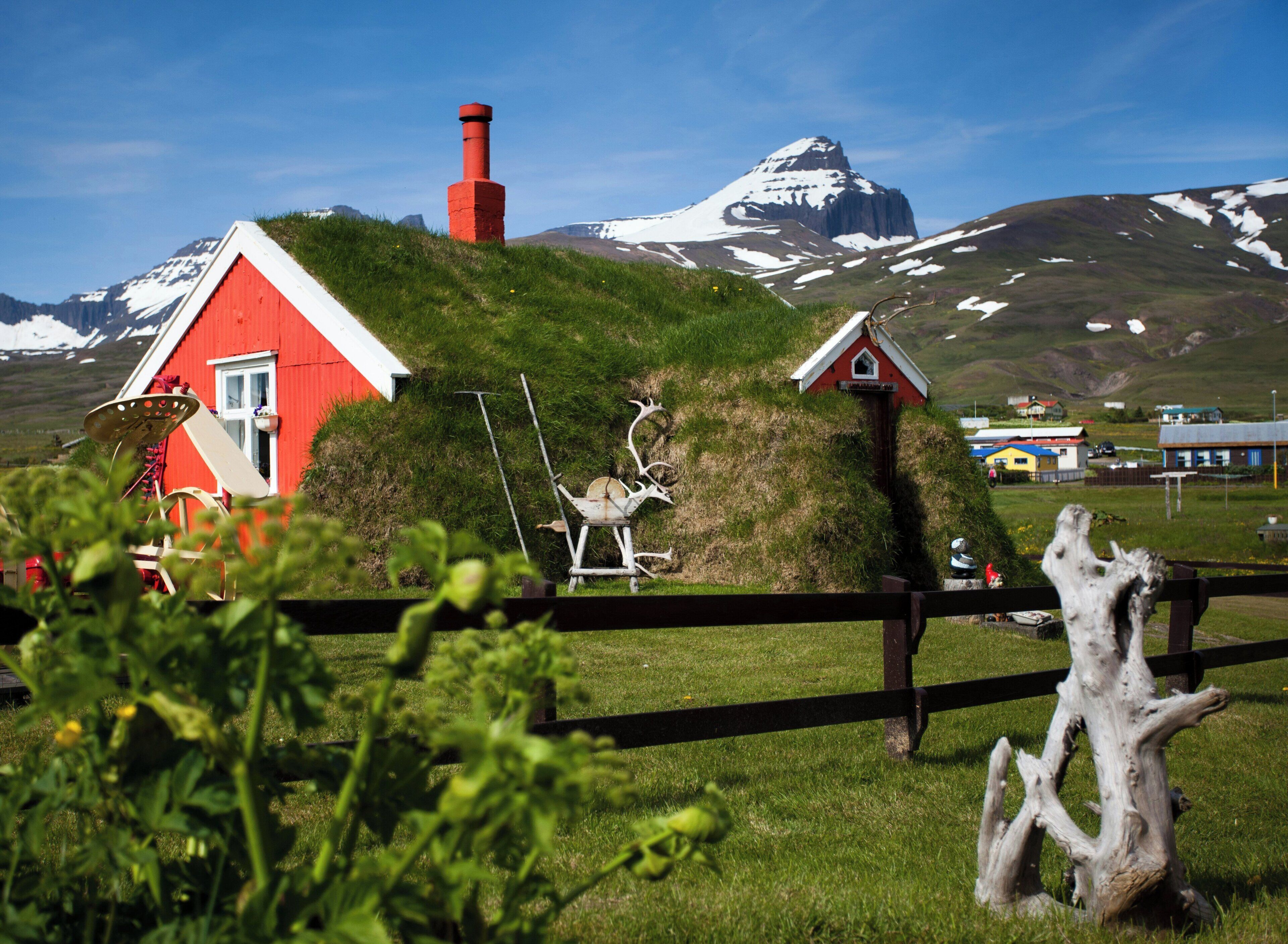 A red house with a white roof, a small shed, and a wooden fence in a grassy field with a mountain in the background.
