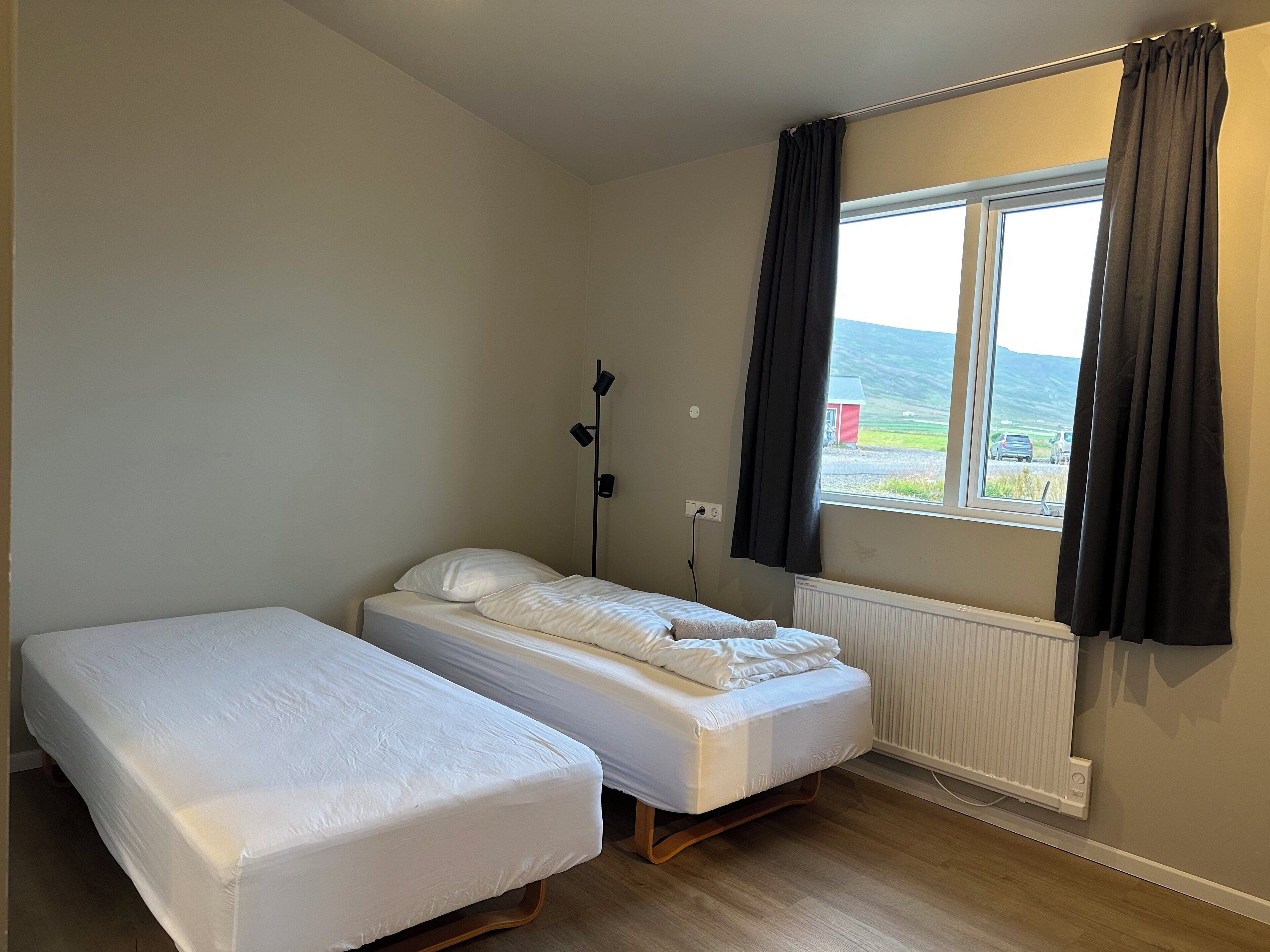 A room with two single beds, a window with curtains, a radiator, and a wall-mounted lamp.
