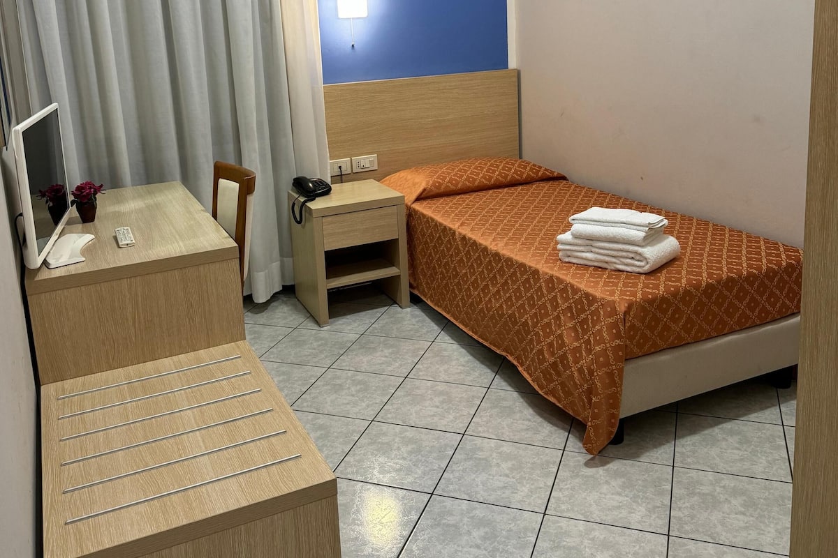 A compact hotel room with a bed, a small desk, a chair, a nightstand with a phone, and a curtain.