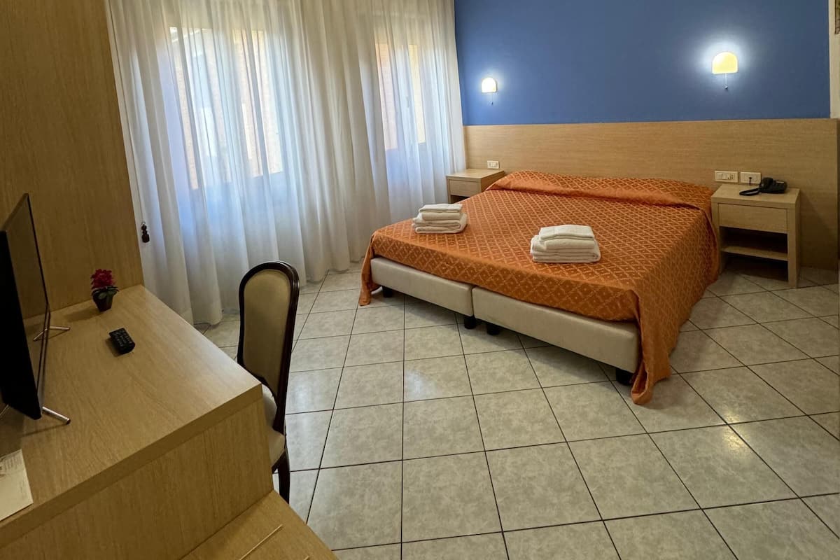 A hotel room with a bed, a desk with a computer, a chair, a nightstand, and a wall-mounted lamp.