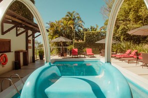 Une piscine avec un toboggan, entourée de chaises longues et de parasols, offrant une vue sur des palmiers et une pergola en bois.
