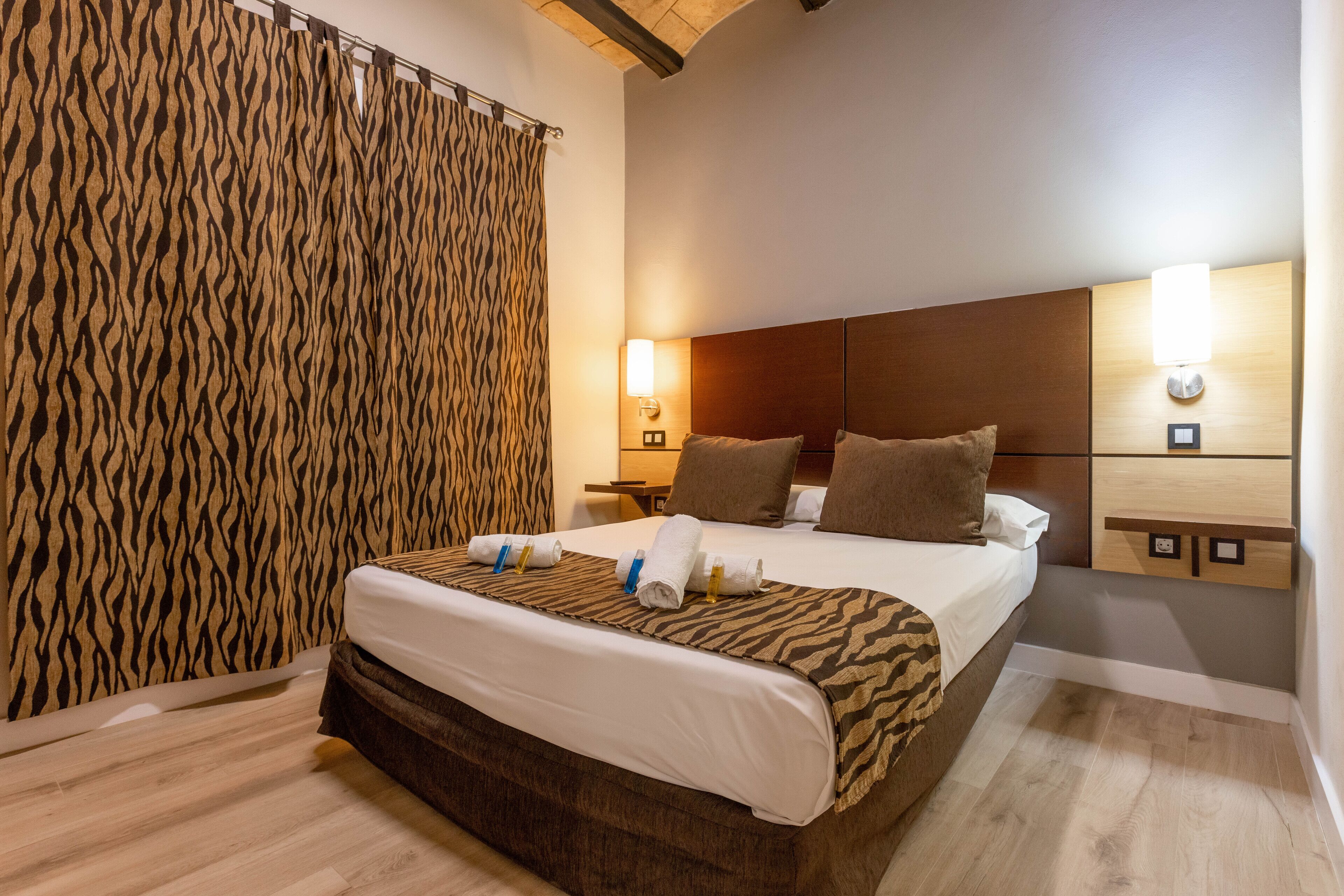 A hotel room with a bed, wooden headboard, two wall-mounted lights, a nightstand, and a patterned curtain.