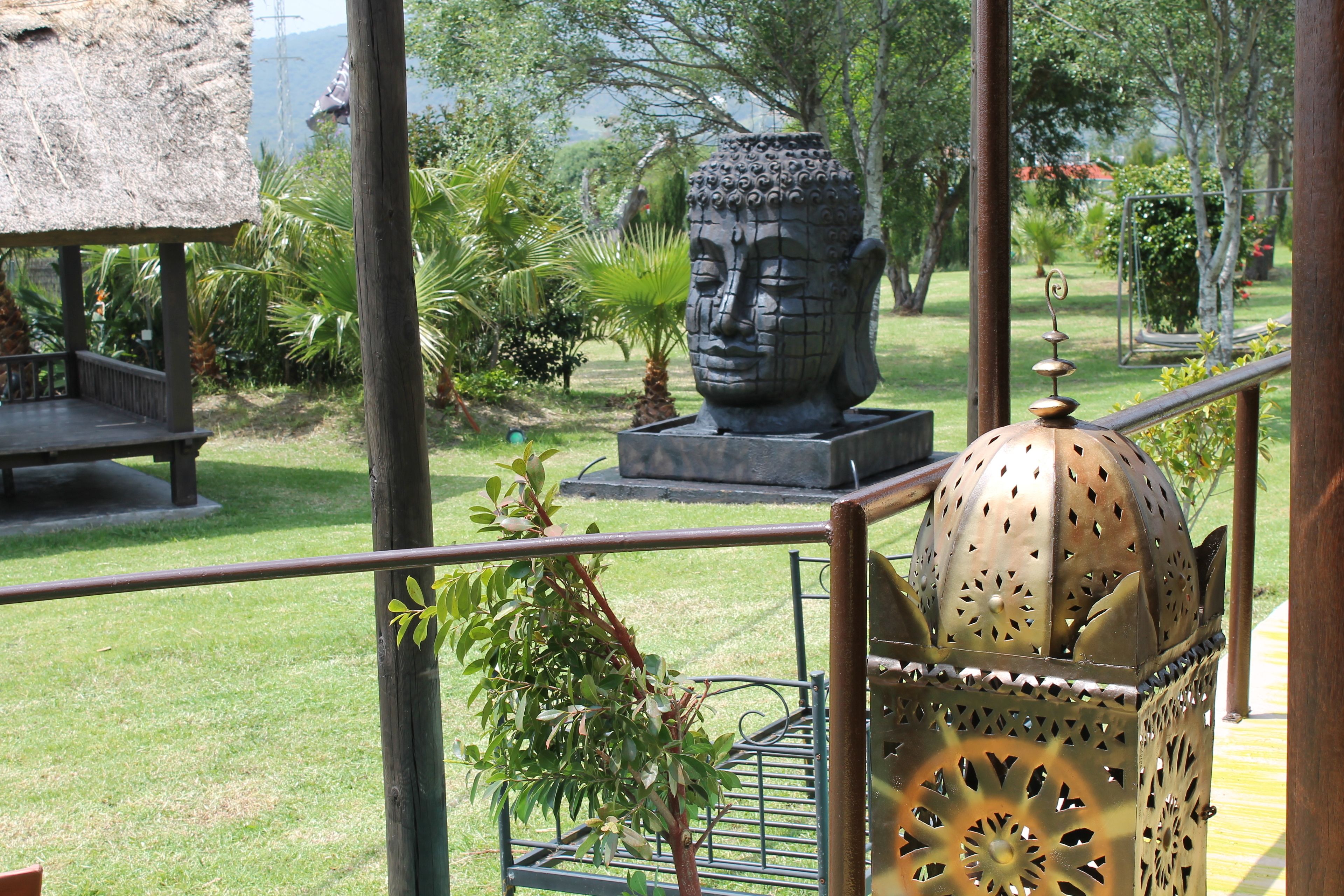 Un jardín con una estatua de Buda, un farol de metal y una estructura con techo de paja.
