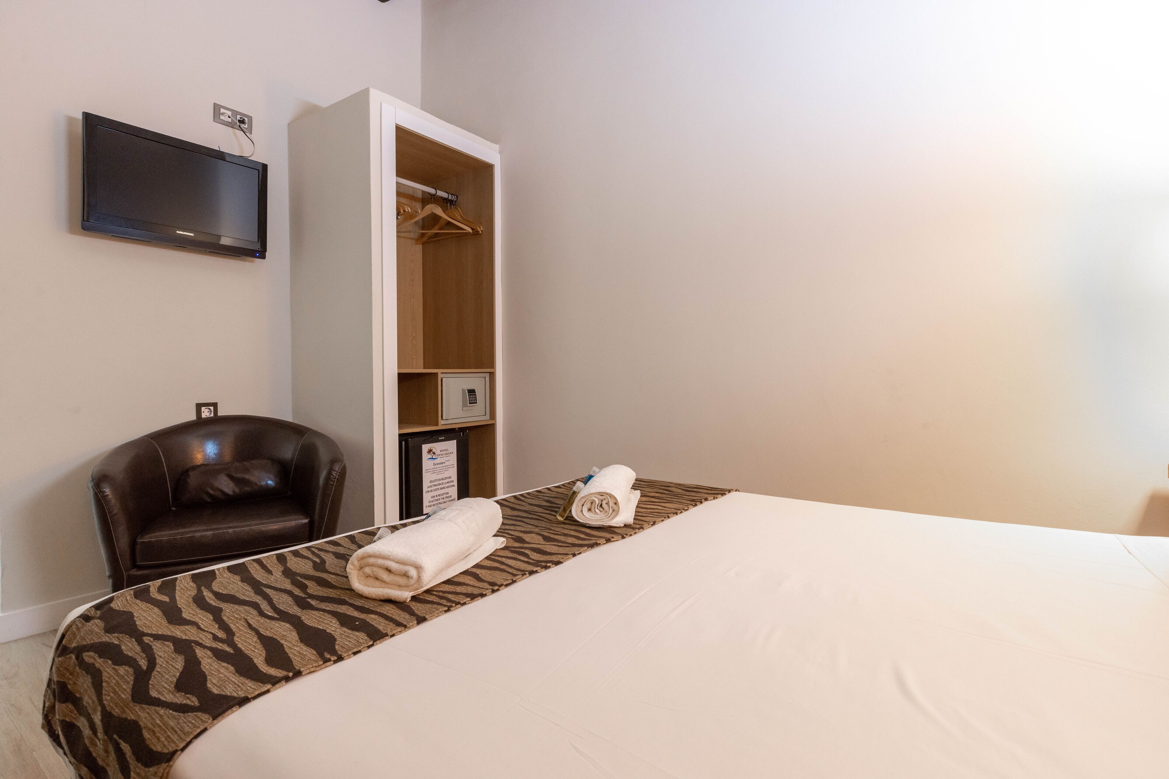 A hotel room with a bed, a TV mounted on the wall, a wardrobe, a leather chair, and a towel on the bed.