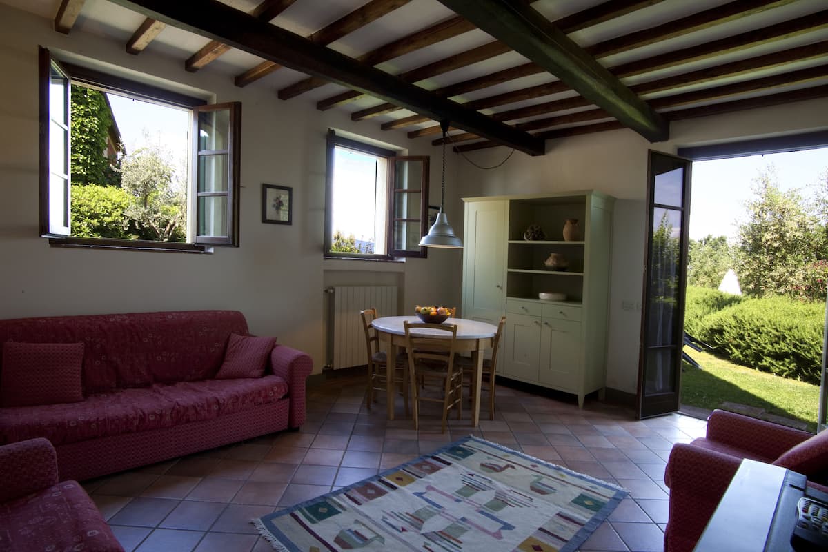 A cozy room with a red sofa, a dining table with chairs, a cabinet with shelves, and a view of greenery outside.
