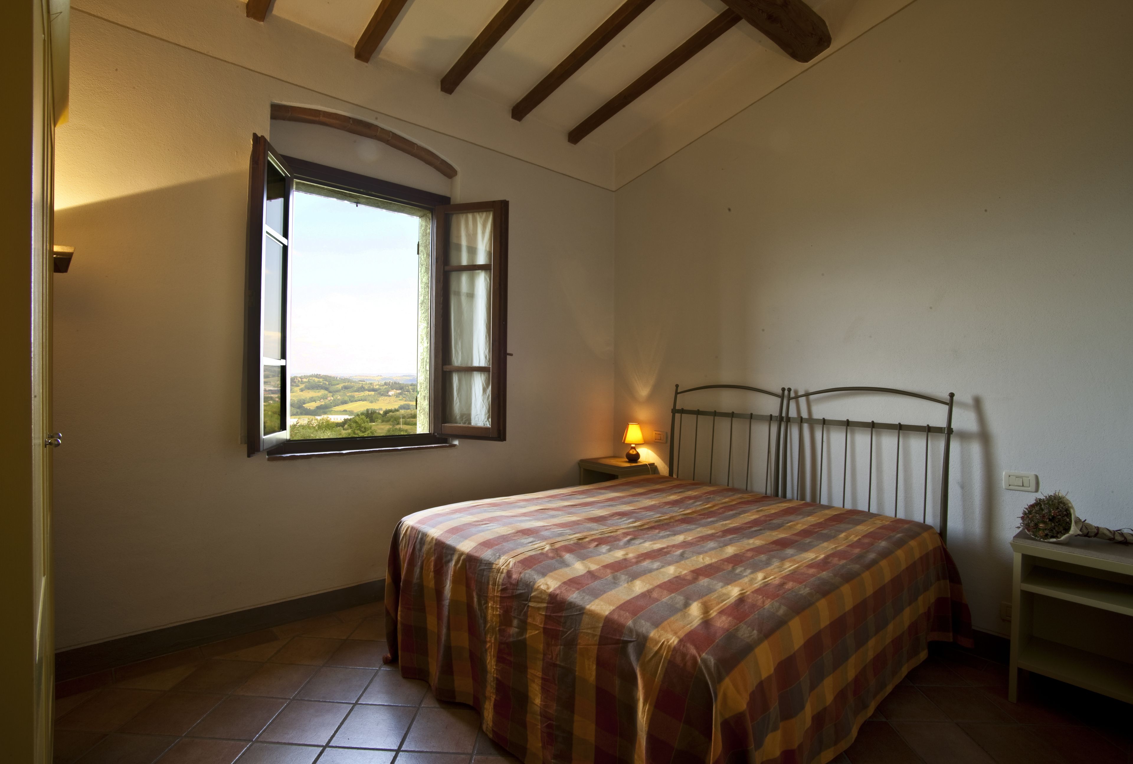 Un dormitorio con cama, ventana con cortinas, mesita de noche con lámpara y cabecera de metal.