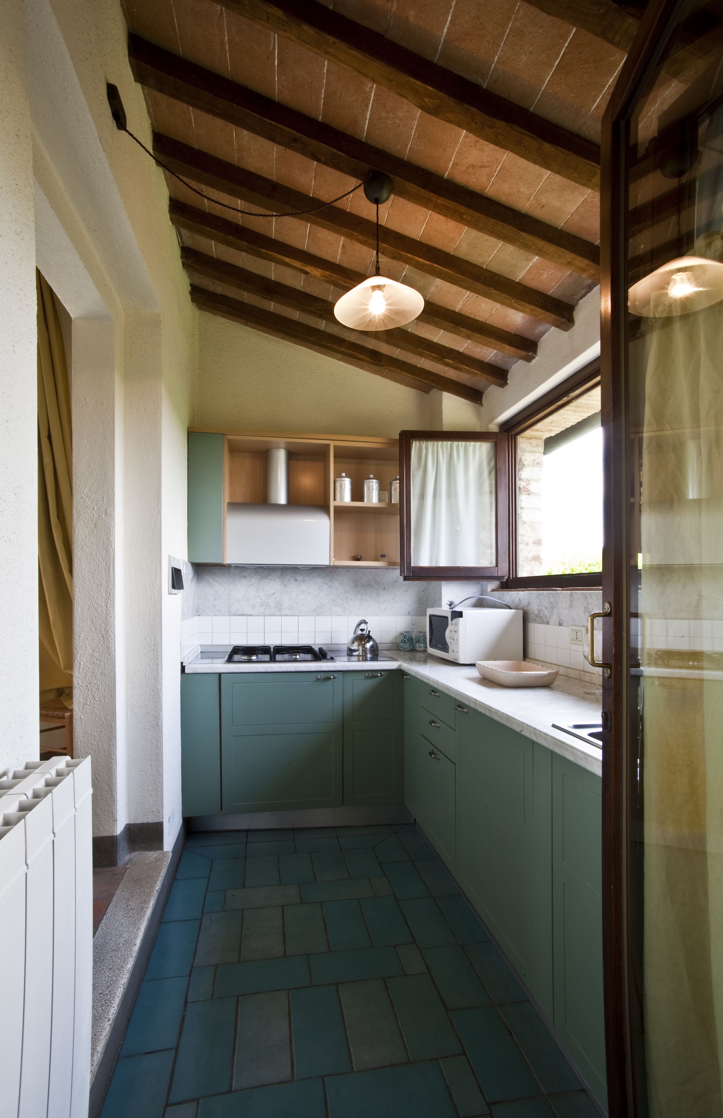 Una cucina compatta con piano cottura a gas, forno a microonde e lavello. Presenta un pavimento piastrellato e travi in legno a soffitto.