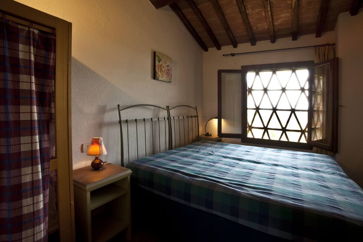 A room with a bed, a nightstand with a lamp, a window with a diamond pattern, and a painting on the wall.