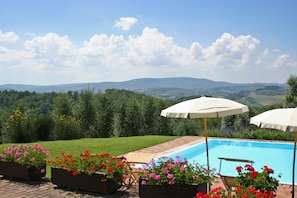 A pool area with a view of distant hills, surrounded by flower beds and umbrellas. - Podere il Pino (Poggibonsi)
