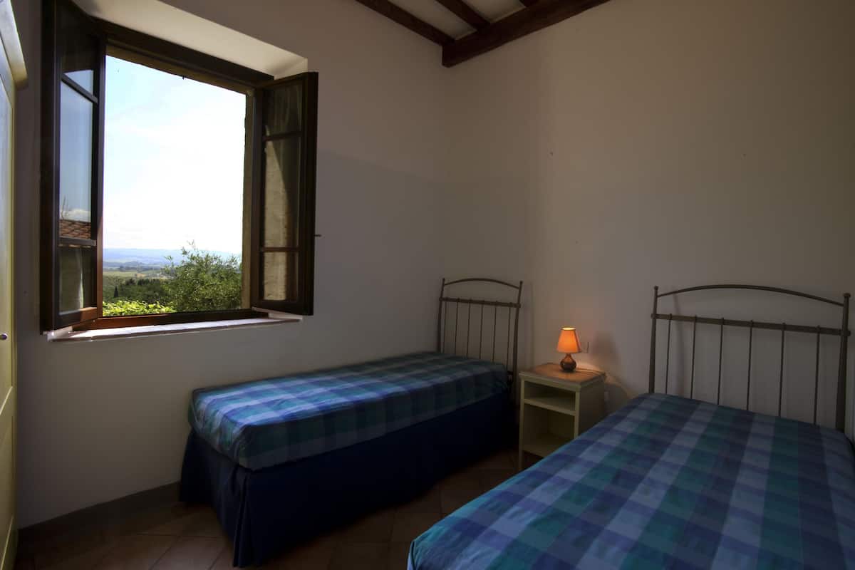 A bedroom with two single beds, a nightstand with a lamp, and a window with a view of greenery.