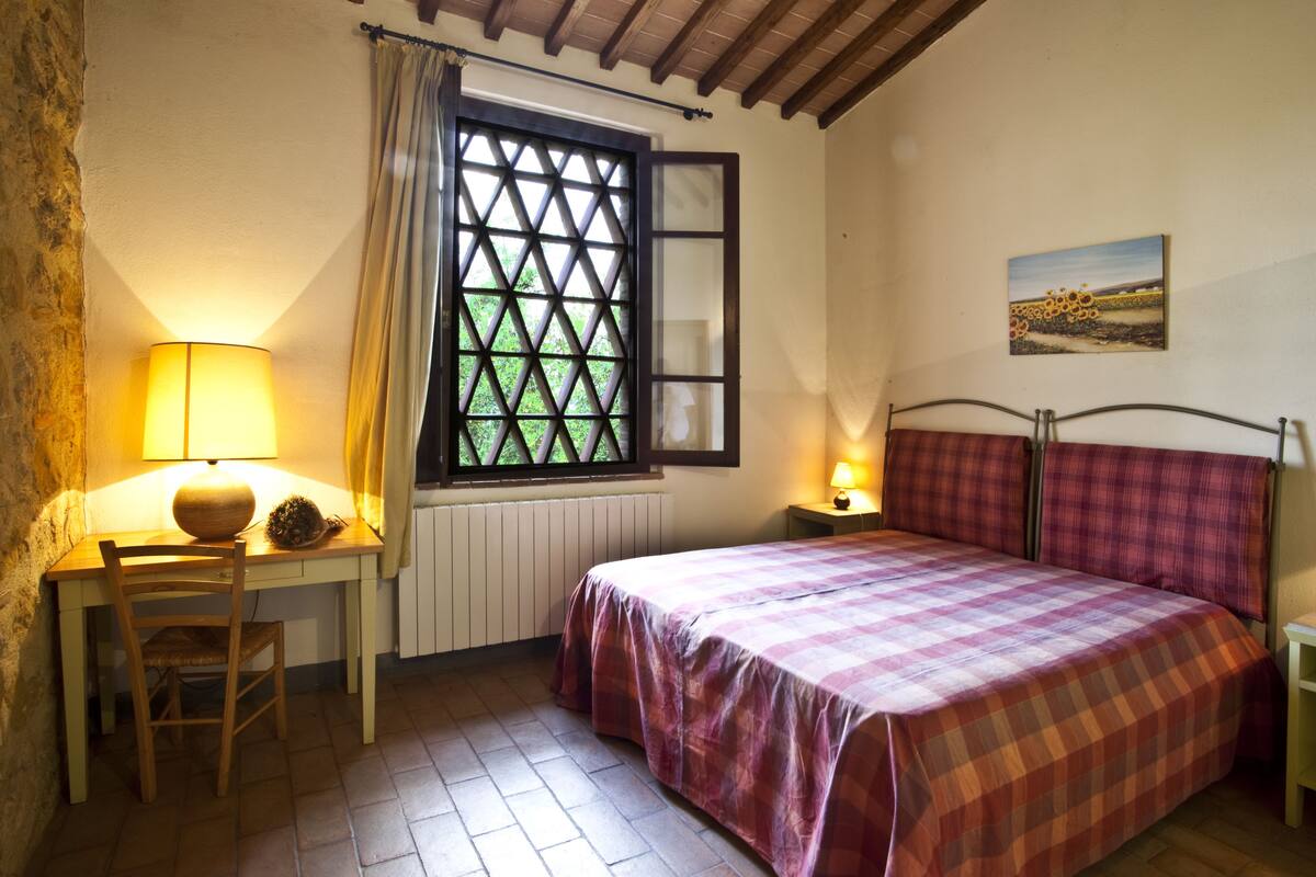 A bedroom with a bed, a desk with a chair, a window with curtains, and a painting on the wall.