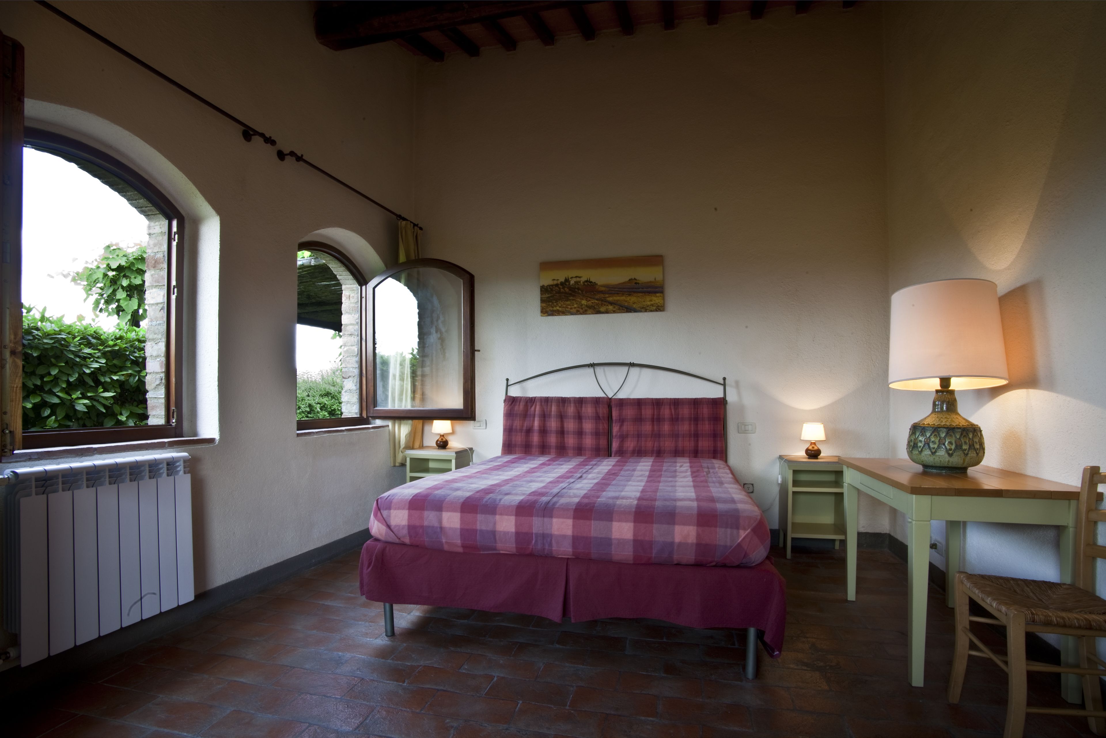 A bedroom with a bed, a desk, a chair, a lamp, a mirror, and a window with a view of greenery.