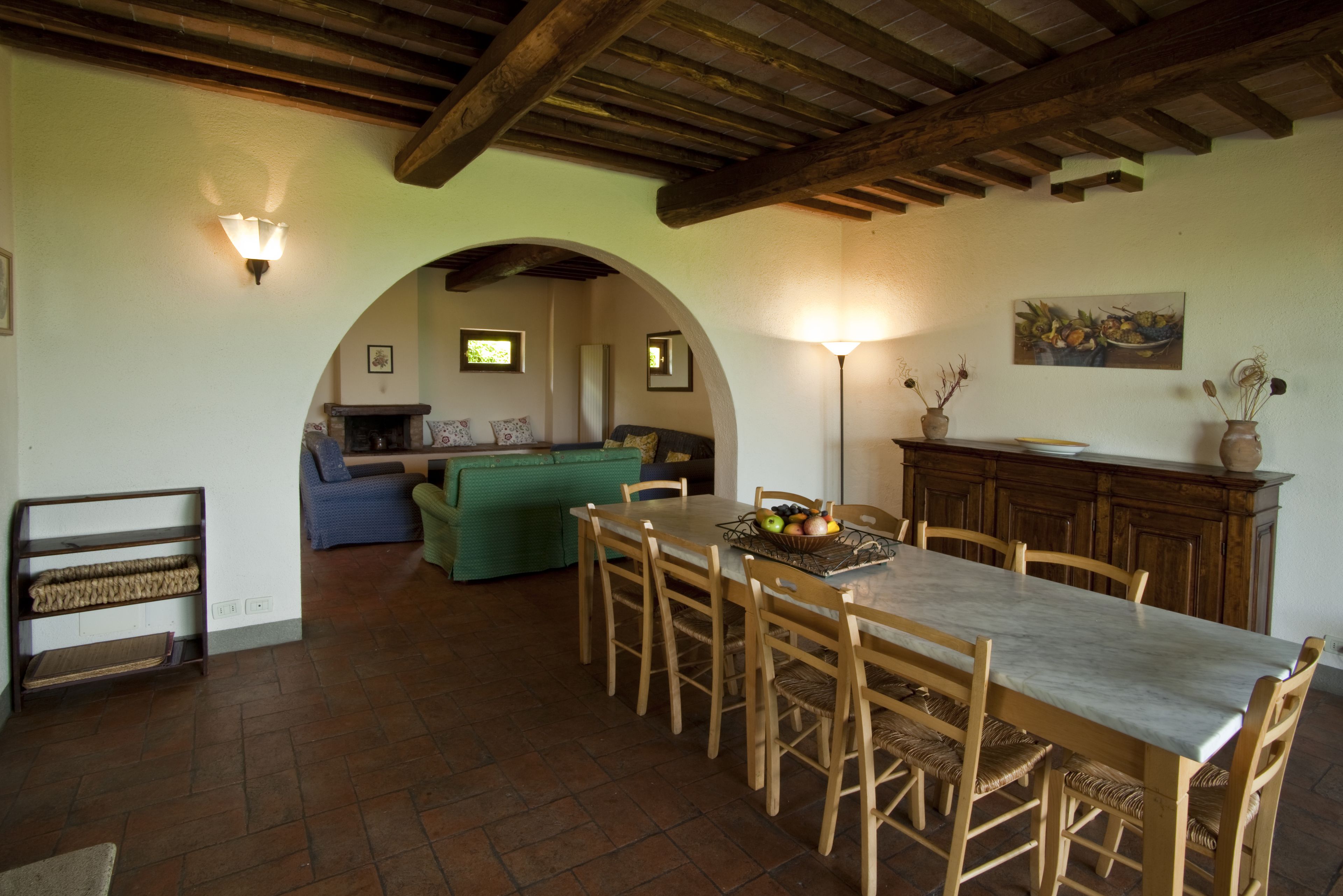 A dining area with a long wooden table, chairs, and a chandelier. There is a green sofa and a fireplace in the background.