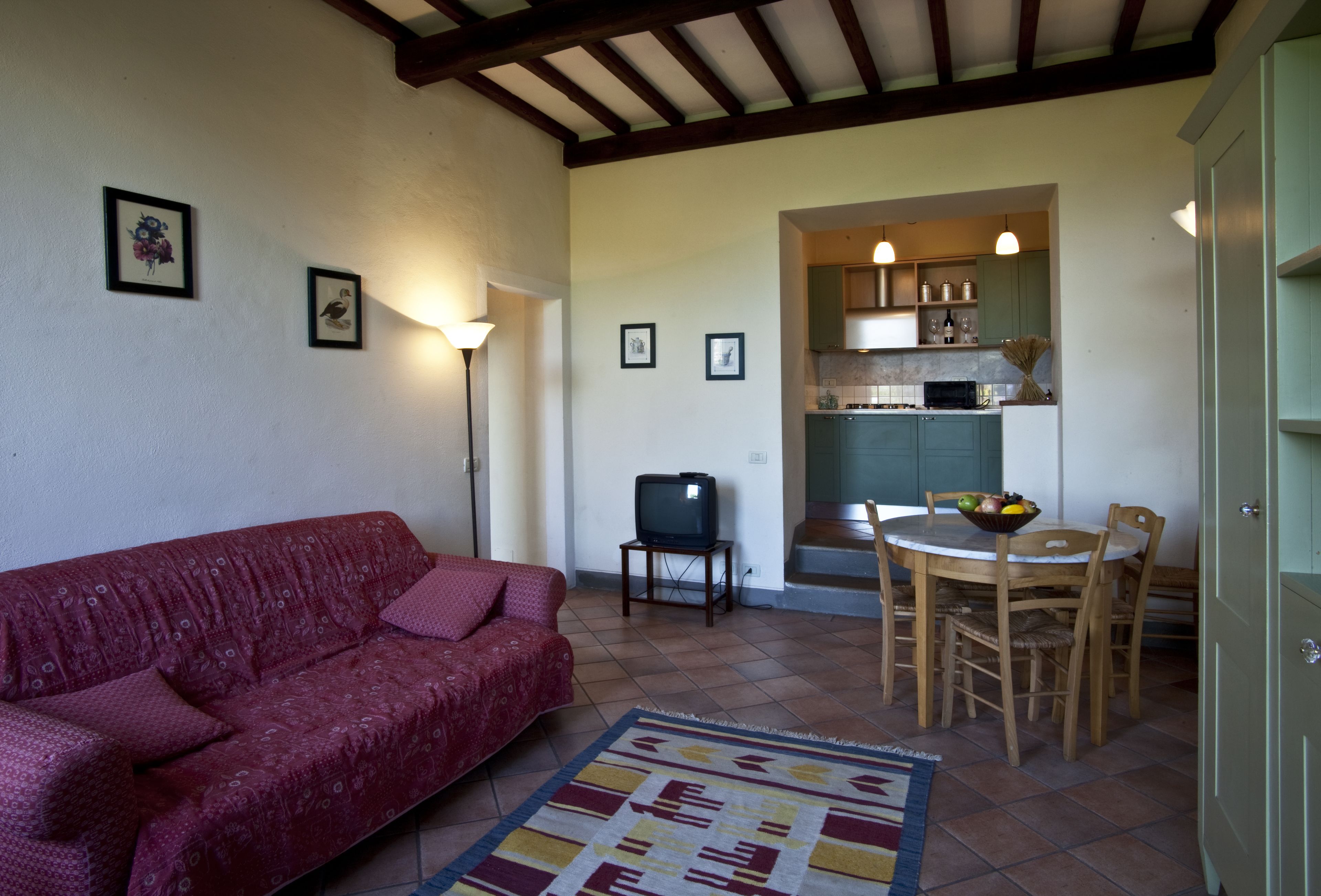 Una sala de estar con un sofá rojo, una mesa de comedor con sillas, un televisor y una zona de cocina con armarios y un microondas.