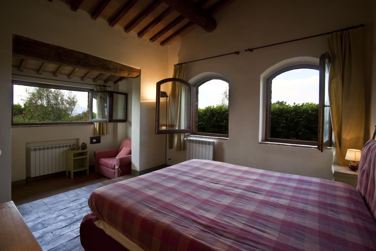 A bedroom with a large bed, a chair, a desk, and two windows.