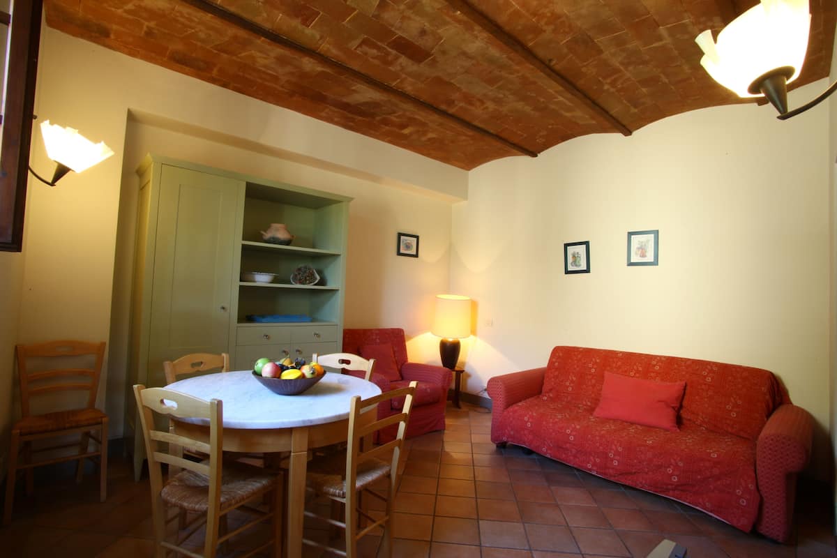 A dining area with a round table, chairs, a bowl of fruit, a red sofa, and a lamp.