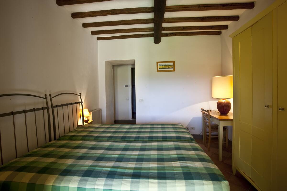 A room with a checkered bedspread, a wooden headboard, a lamp on a table, and a yellow wardrobe.