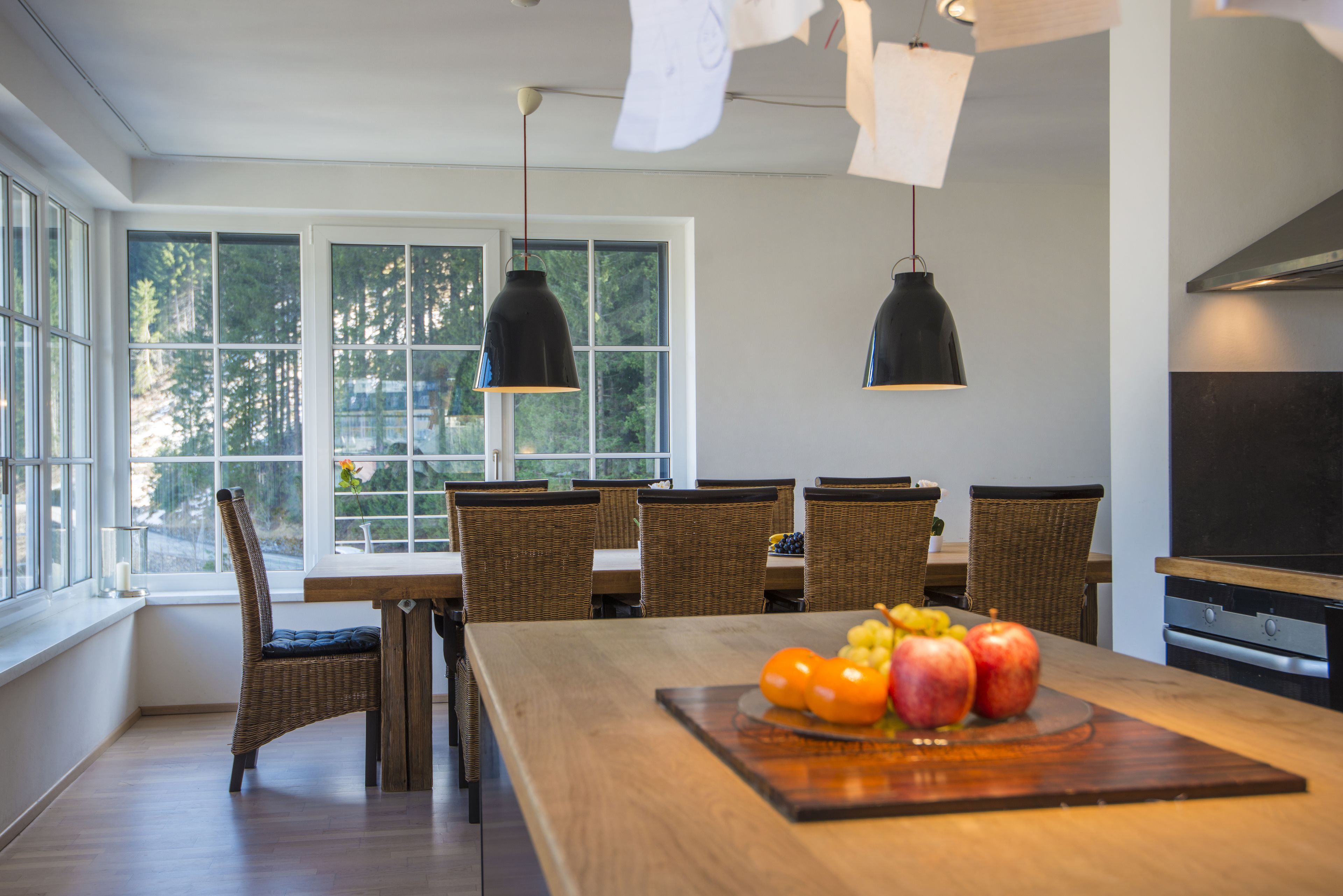 A modern kitchen with a wooden dining table, wicker chairs, and pendant lights.