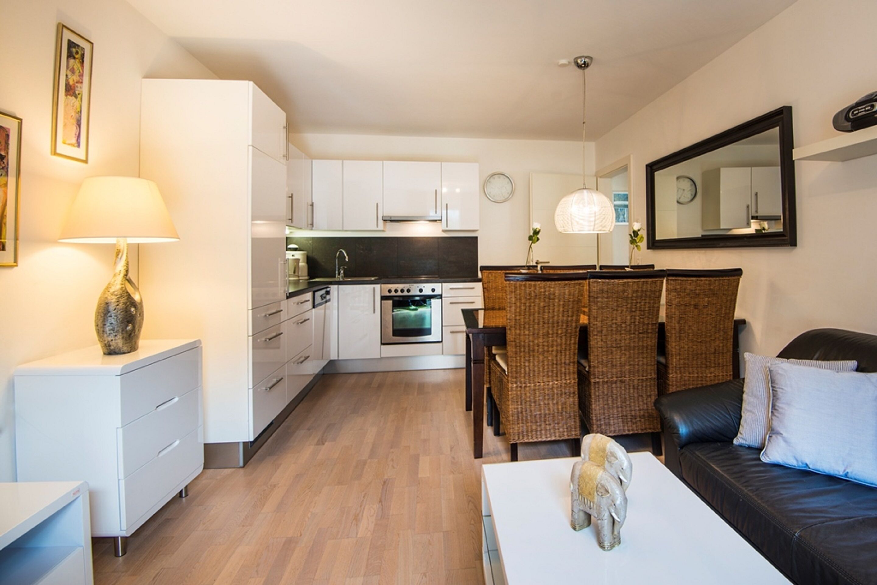 A modern kitchen with white cabinets, a dining area with wicker furniture, and a living room with a black sofa and a white coffee table.