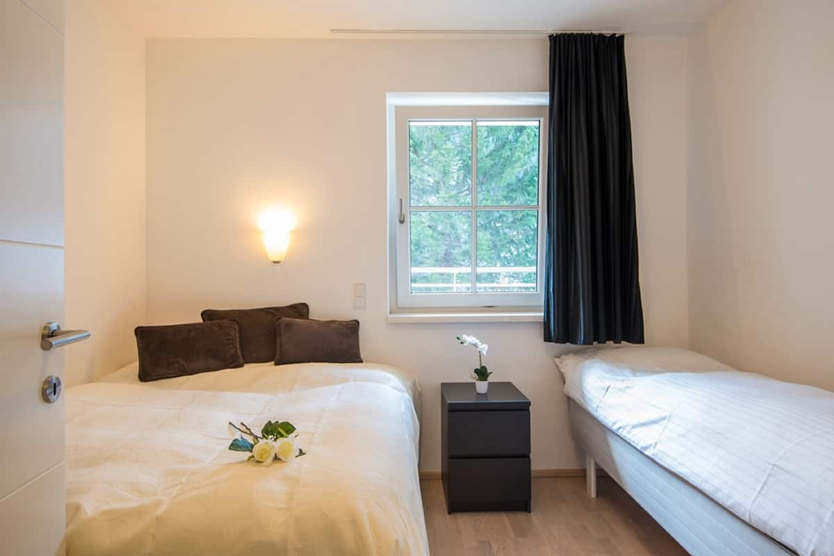 Un dormitorio con dos camas, una ventana con cortinas, una mesita de noche con una planta y una lámpara fijada a la pared.