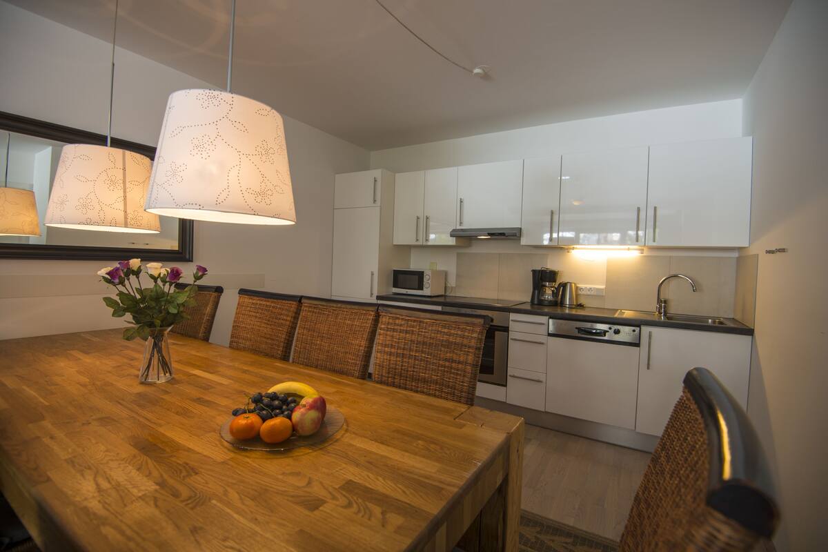 Una cocina moderna con armarios blancos, un microondas y un lavamanos. Hay una mesa de comedor con un frutero y un florero.