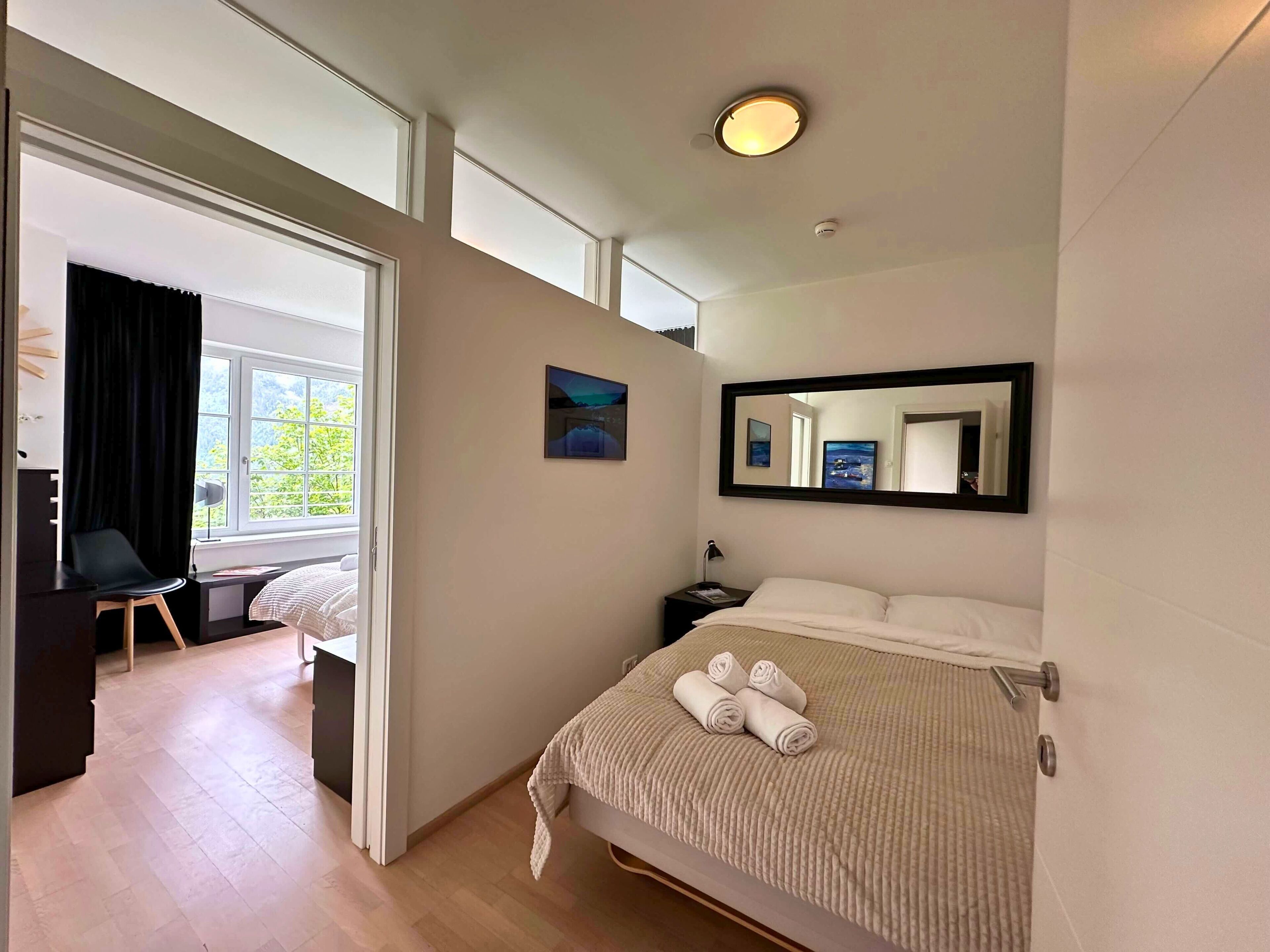 Habitación de hotel con cama, escritorio, silla, ventana con cortinas, un cuadro, un espejo y una lámpara en el techo.