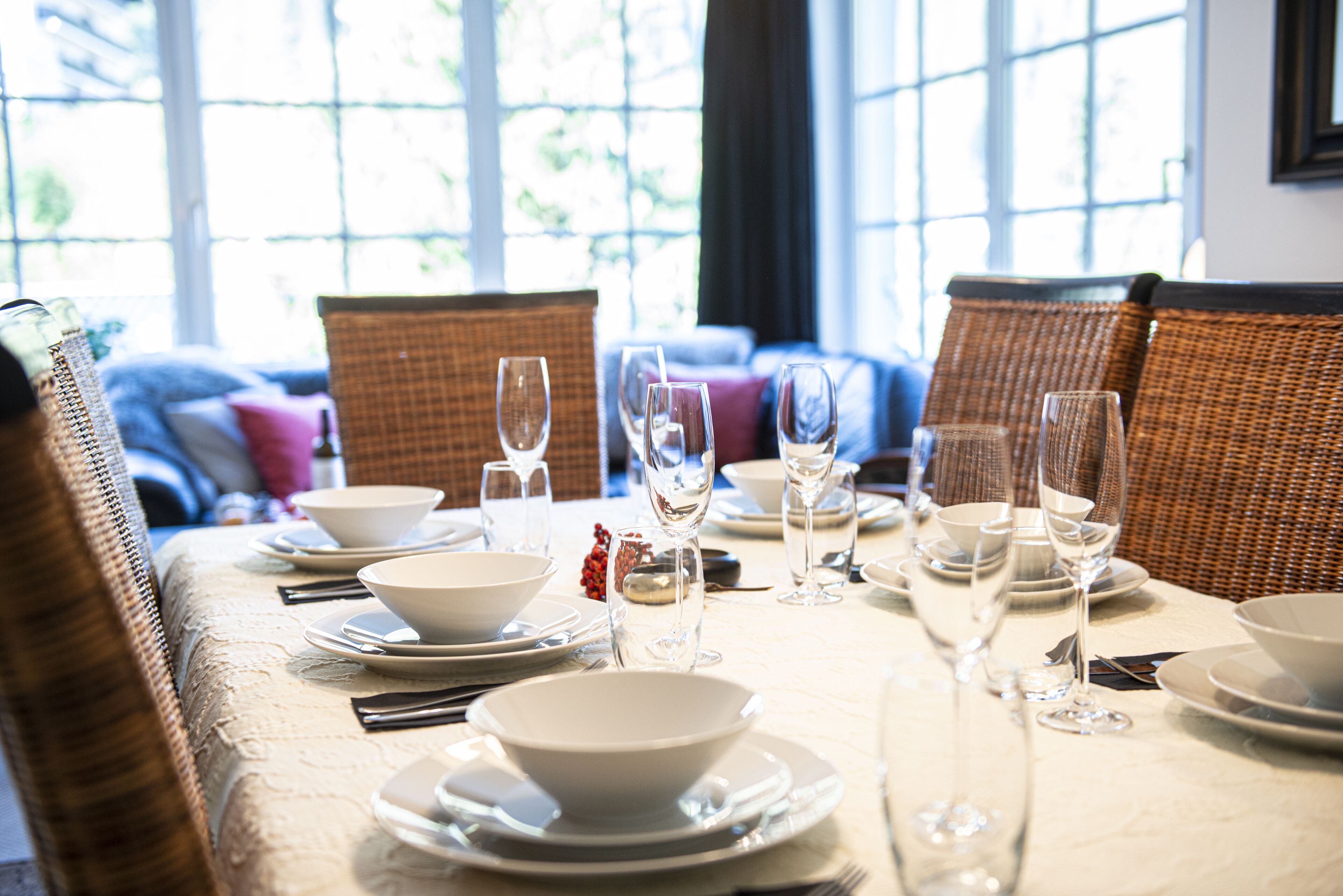 Une table à manger dressée avec de l’assiette blanche, des bols et des verres à vin, entourée de chaises en osier.