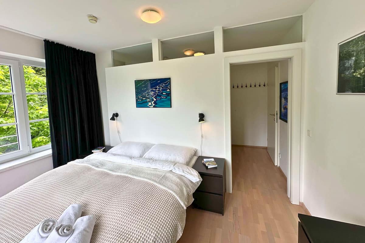 Un dormitorio con cama, mesitas de noche, lámpara de techo, ventana con cortinas, un cuadro en la pared y un pasillo que conduce a otra habitación.