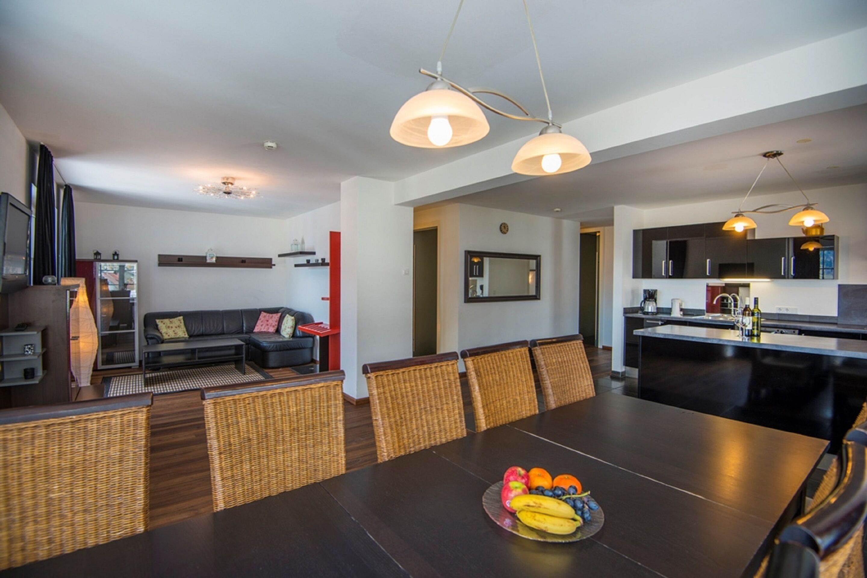 A modern kitchen with dark countertops, a dining area with a wooden table, wicker chairs, and a fruit bowl.