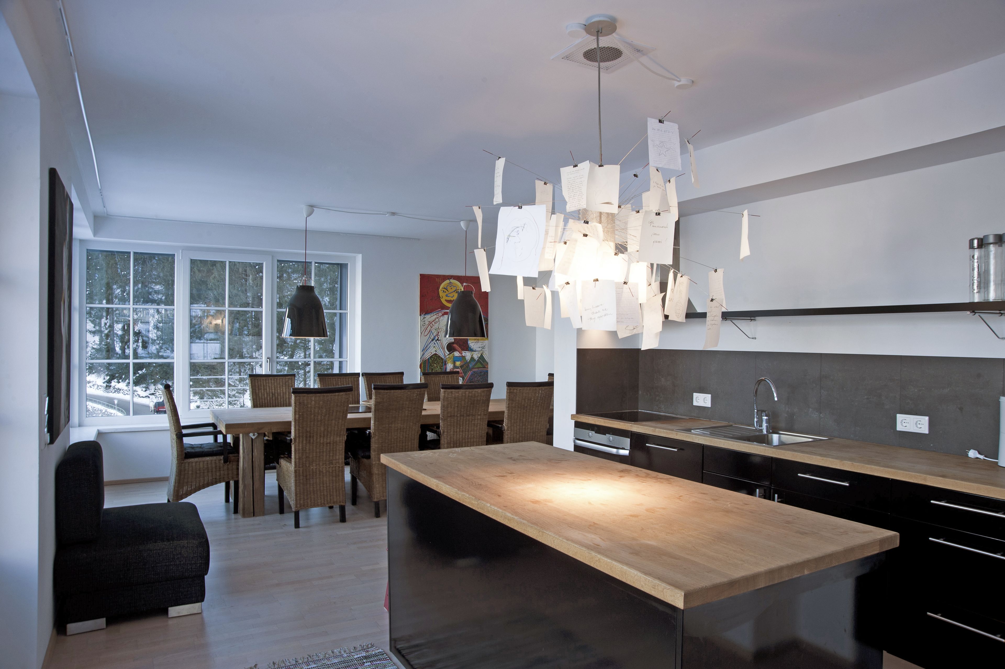 Une cuisine moderne dotée d’un îlot central, d’un coin repas avec des chaises en osier et d’une grande fenêtre offrant une vue sur les arbres.