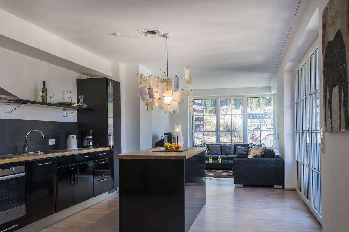 Una cocina moderna con una isla central, un comedor con sofá y grandes ventanales con vistas a la ciudad.