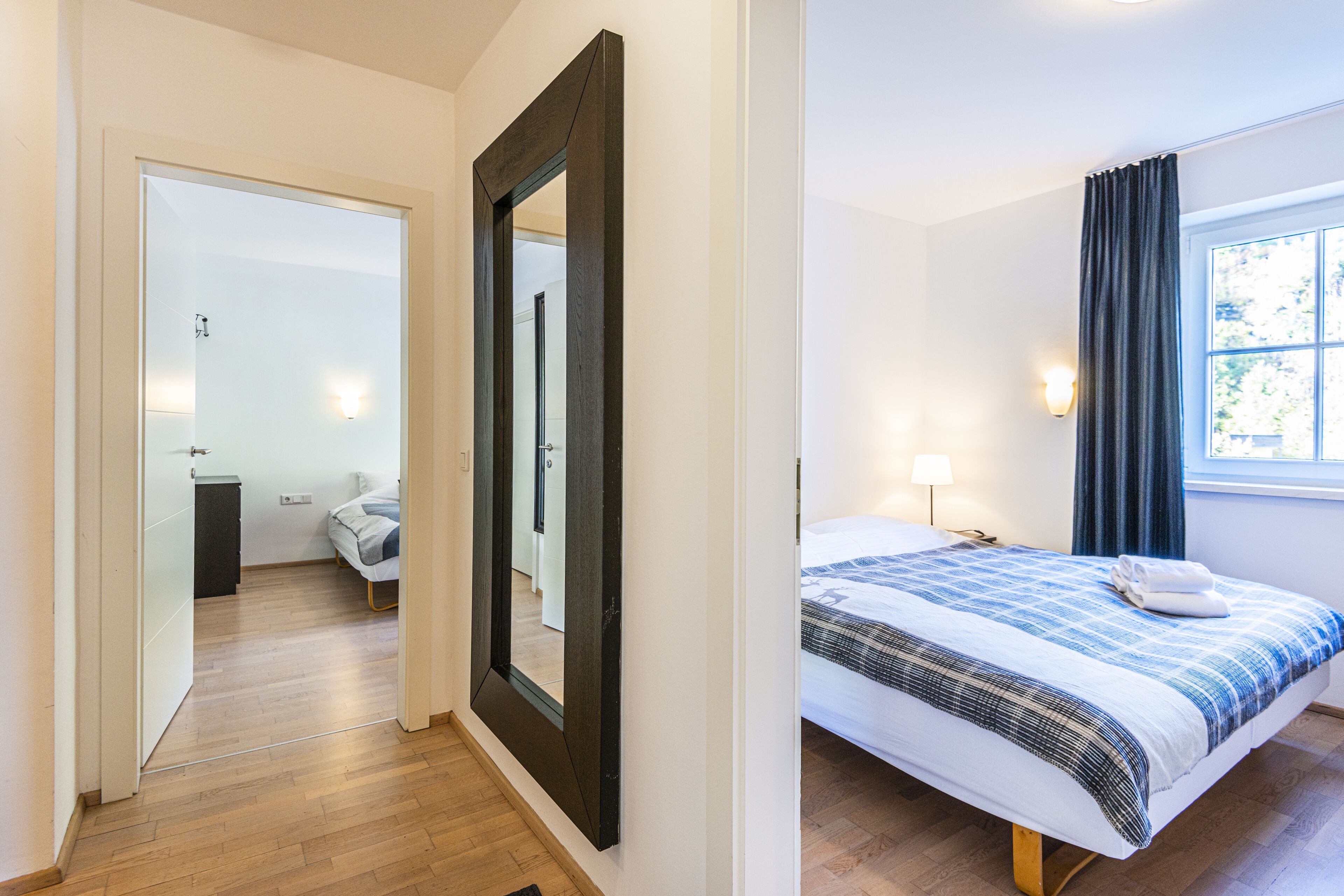 Une chambre d’hôtel avec un plancher en bois, un grand miroir, un lit recouvert d’une courtepointe à carreaux bleus et blancs, et une fenêtre avec des rideaux.