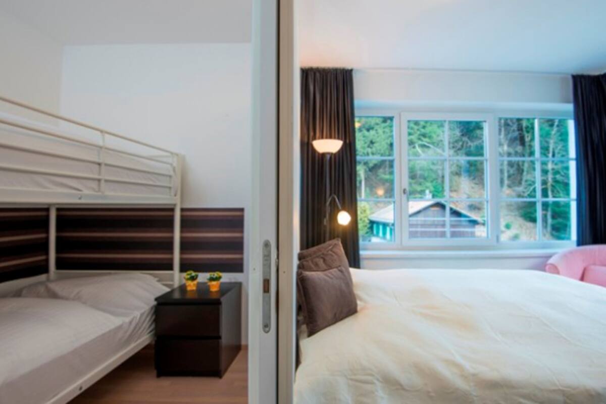 Un dormitorio con una litera, una cama individual, una mesita de noche con lámpara y una ventana con cortinas.
