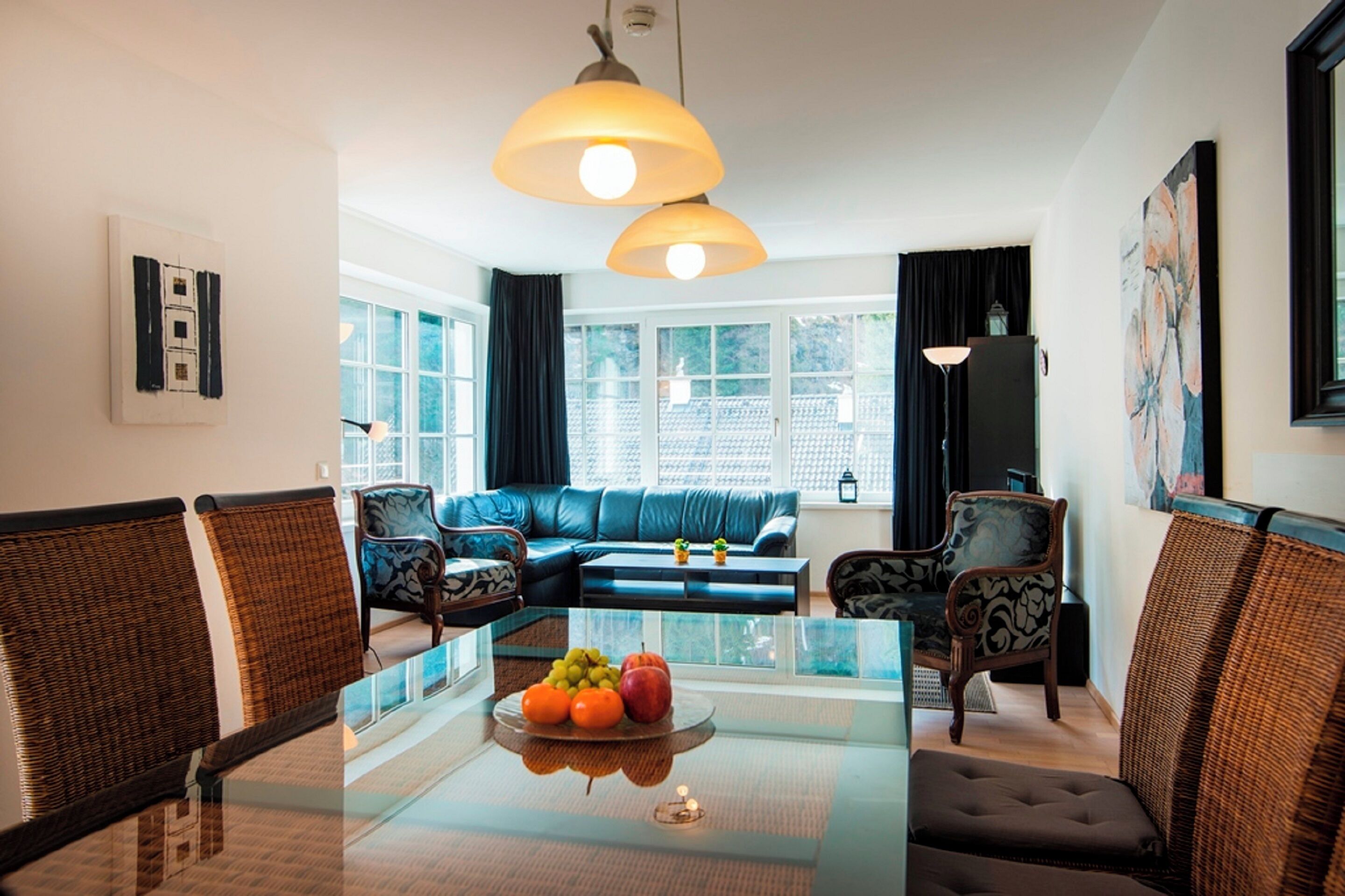 A modern living room with a glass dining table, wicker chairs, a blue sofa, and a large window with curtains.