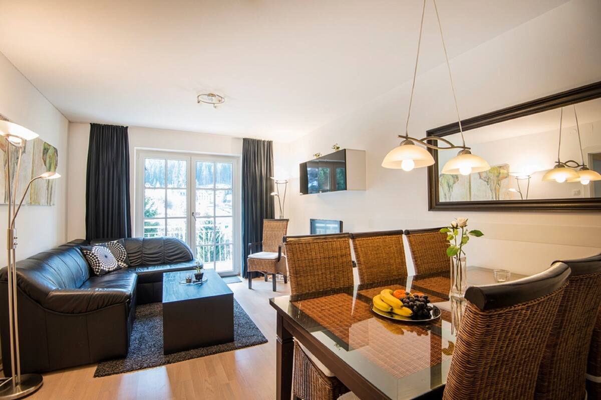 Una sala de estar moderna con un sofá, una mesa de comedor y sillas. Hay un ventanal con cortinas, un televisor de pantalla plana y un espejo sobre la mesa de comedor.