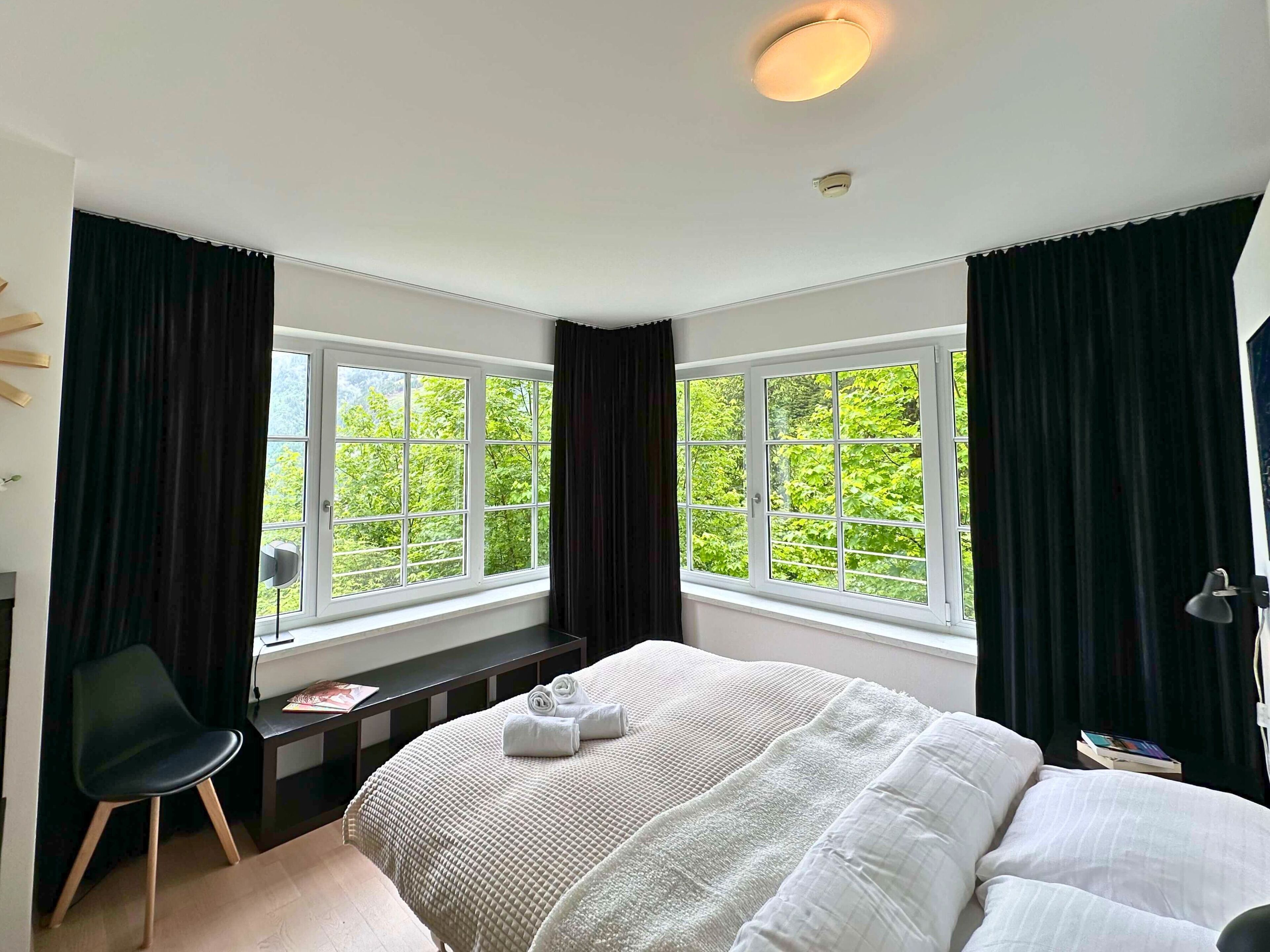 A bedroom with a large bed, two windows with curtains, a desk, and a chair.