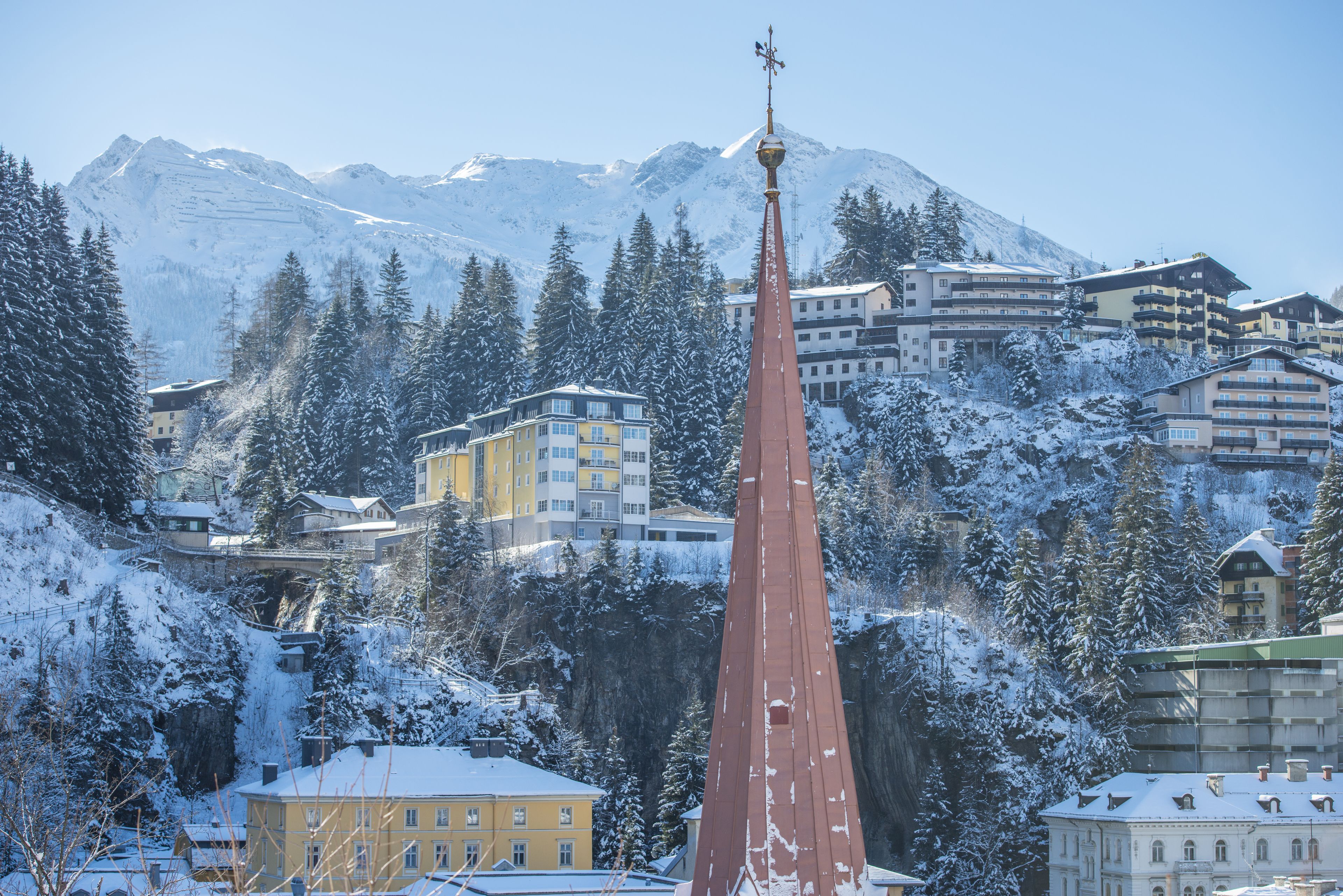 A snow-covered mountain town with a prominent church spire, residential buildings, and a clear view of the mountains in the background.