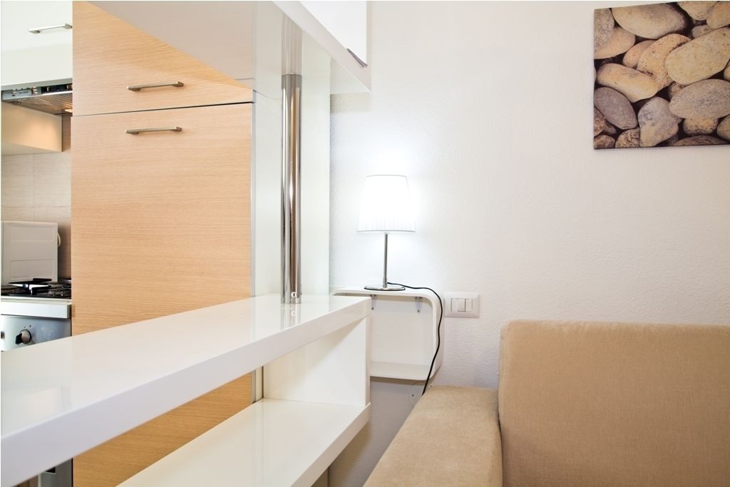 A modern kitchen with white cabinetry, a built-in stove, and a countertop. There is a beige sofa in the living area and a lamp on the wall.