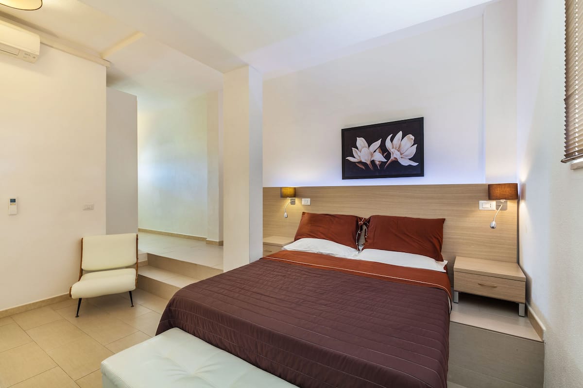 A modern hotel room with a large bed, a chair, a nightstand, and a wall-mounted artwork.