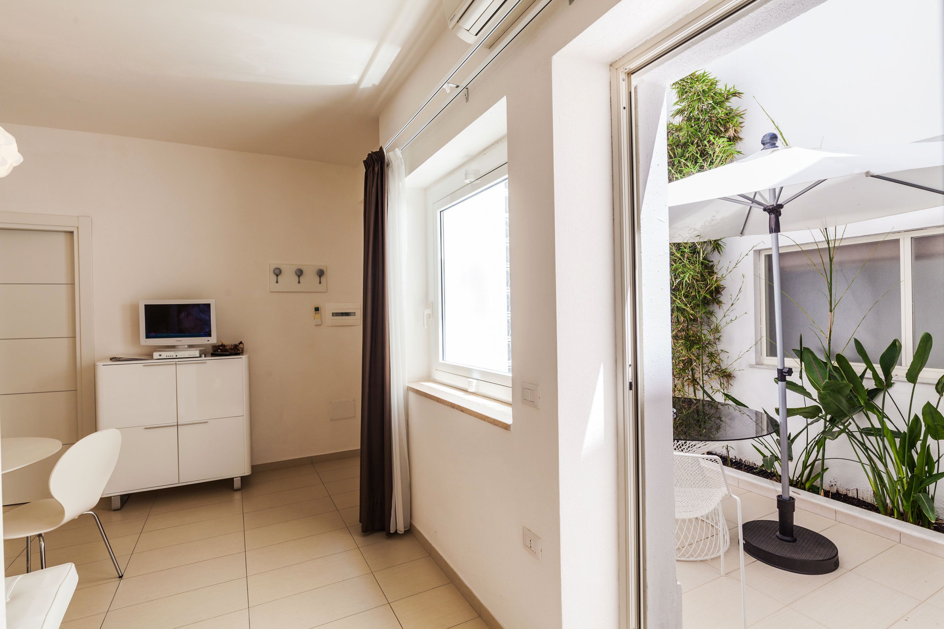 Una habitación moderna con televisor en un mueble, una zona de comedor con sillas, un ventanal con cortinas y un balcón con mesa y sombrilla.