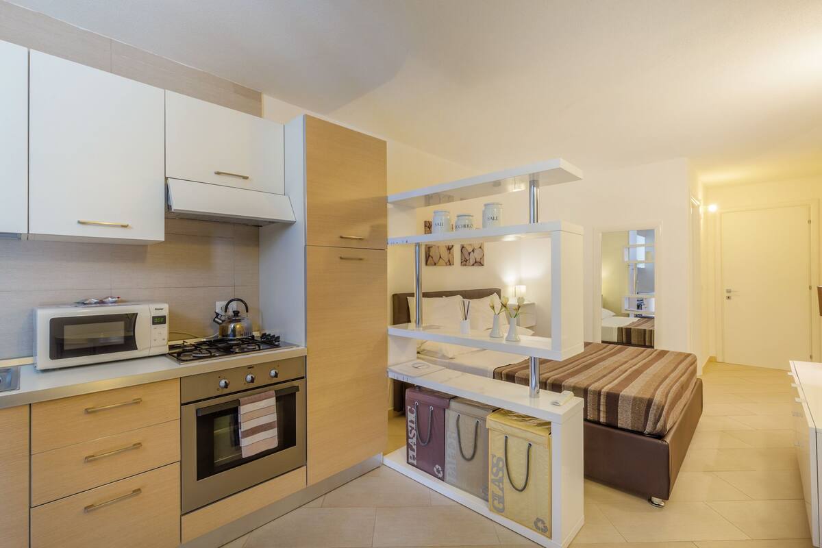 A compact kitchen with a microwave, stove, and oven. There is a bed with a striped blanket and a bedside table with a lamp.